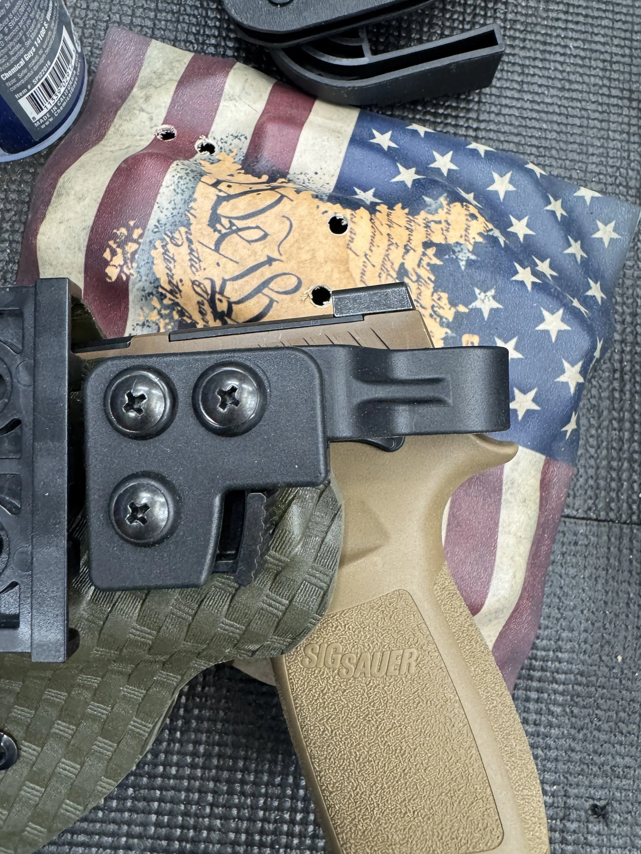 A tattered American flag with a skull logo, a Glock pistol, a gun mount, a can of cleaning product, and a leatherman multi-tool on a textured black surface.