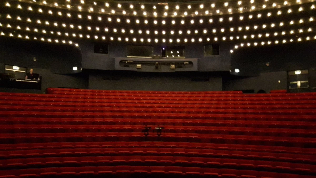 Empty theater auditorium with red seats and a stage, illuminated by ceiling lights.