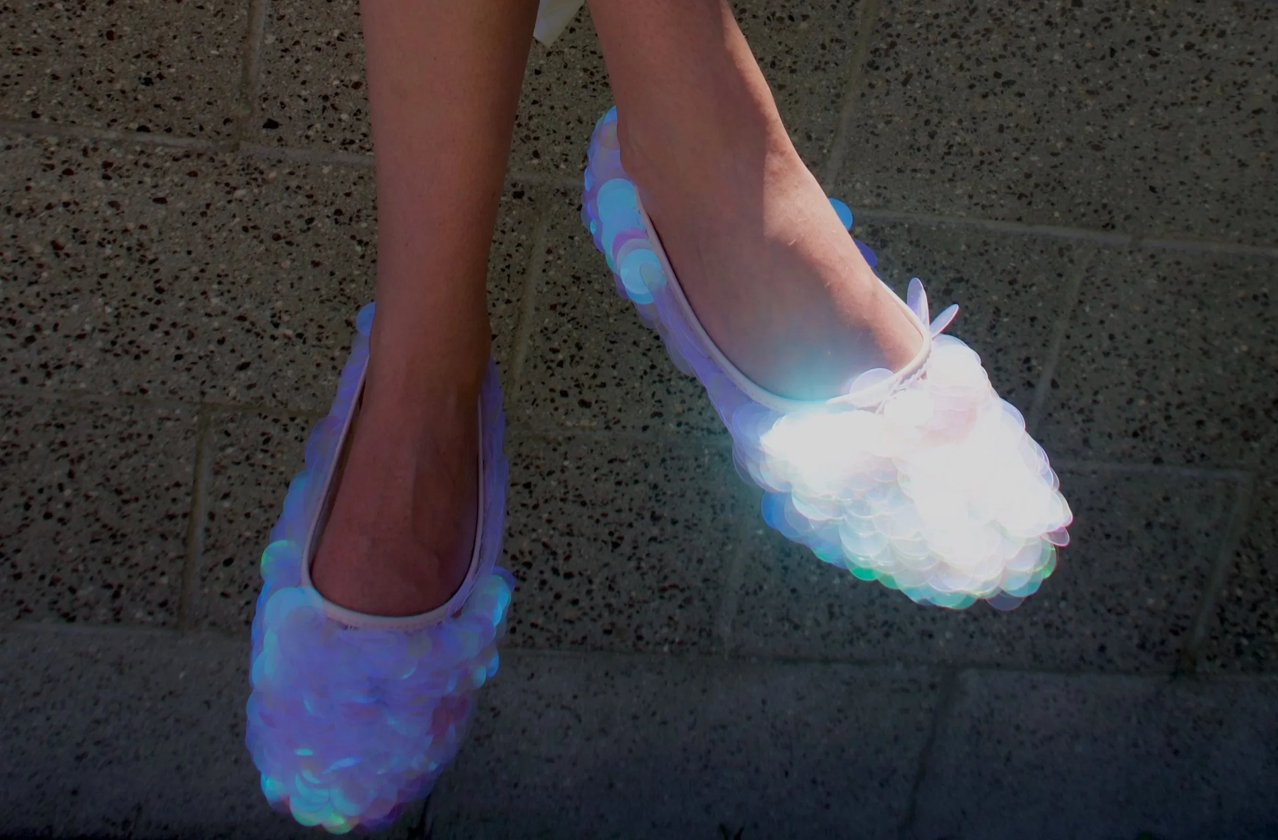 A person's feet wearing iridescent shoes covered in circular, holographic sequins, standing on a tiled floor.