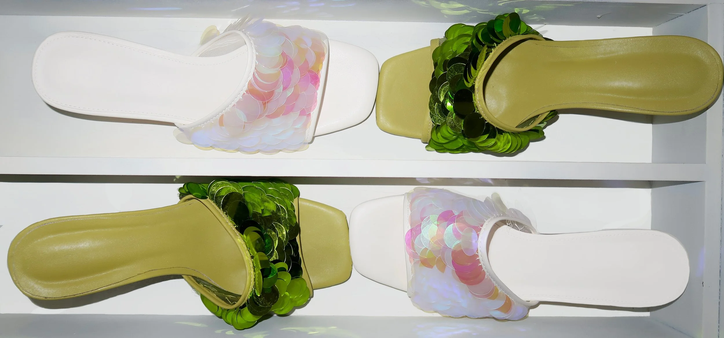 Pair of slide sandals, one white and one yellow, decorated with shiny sequins, arranged on a white shelf.