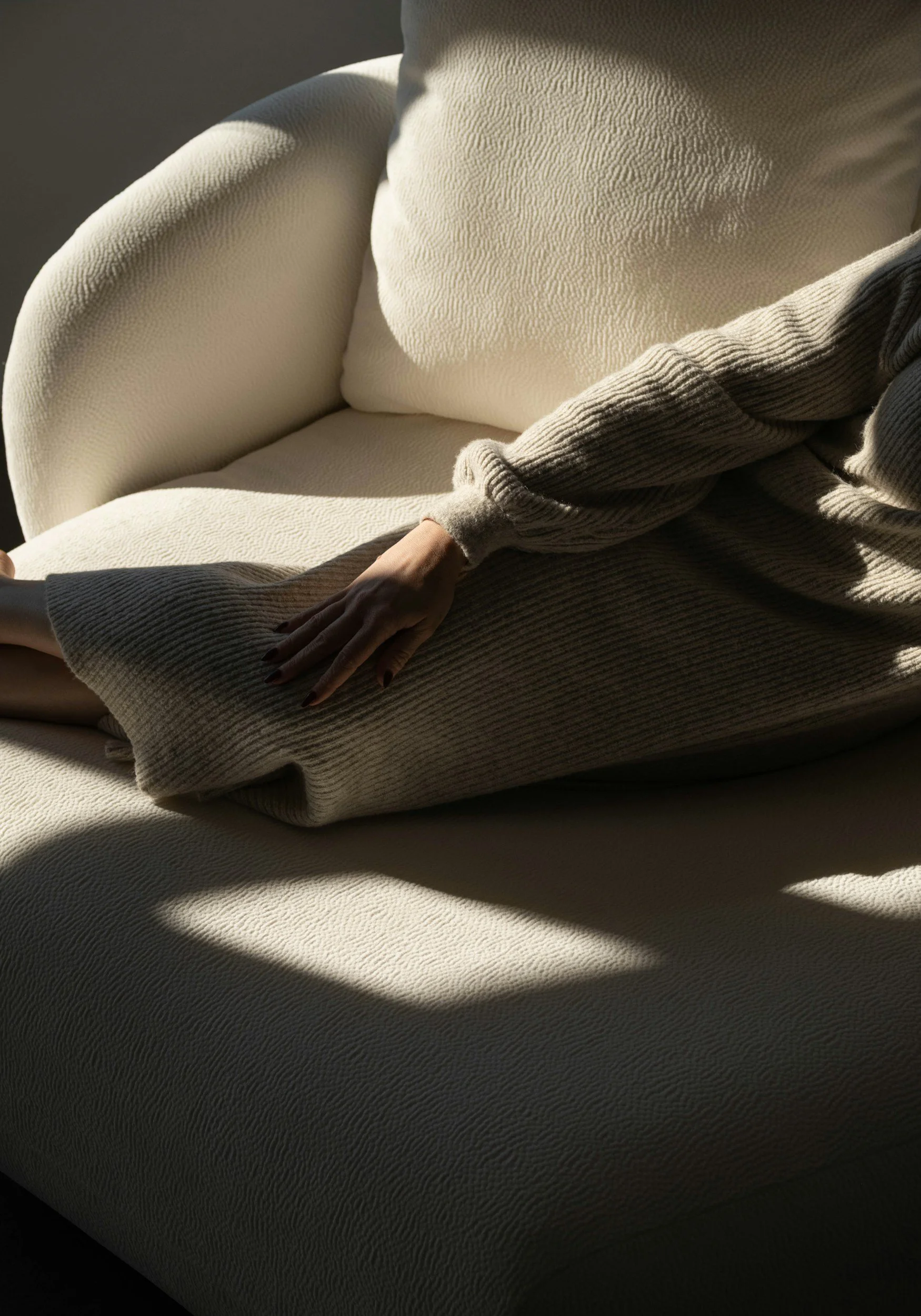 A person sitting on an off-white textured sofa, wearing a beige knit dress and a gray knit sweater, with their hand resting on their knee, illuminated by soft natural light.