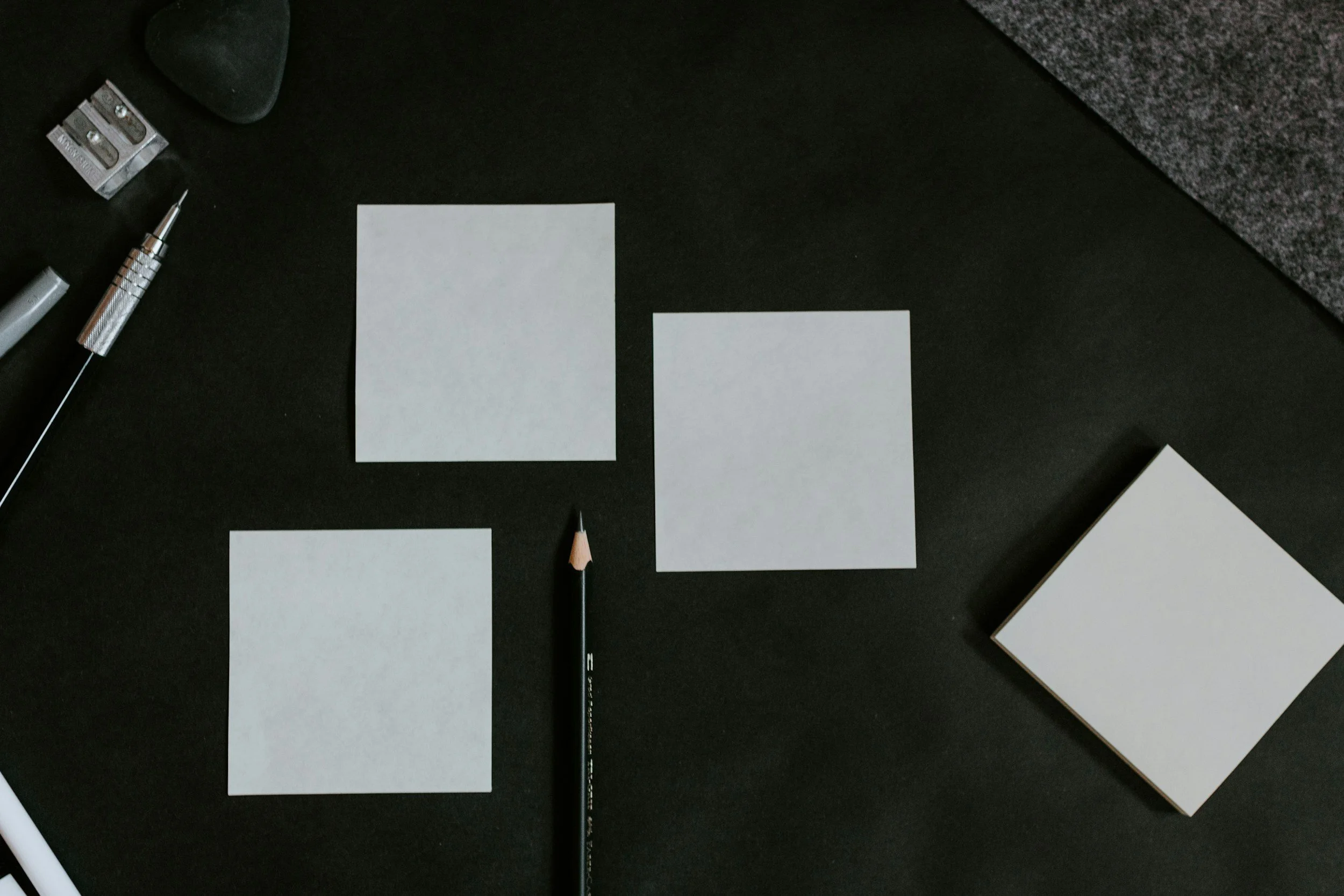 Three blank white square sticky notes on black surface, with a black pencil, a silver mechanical pencil, a gray eraser, and a stack of blank white sticky notes.