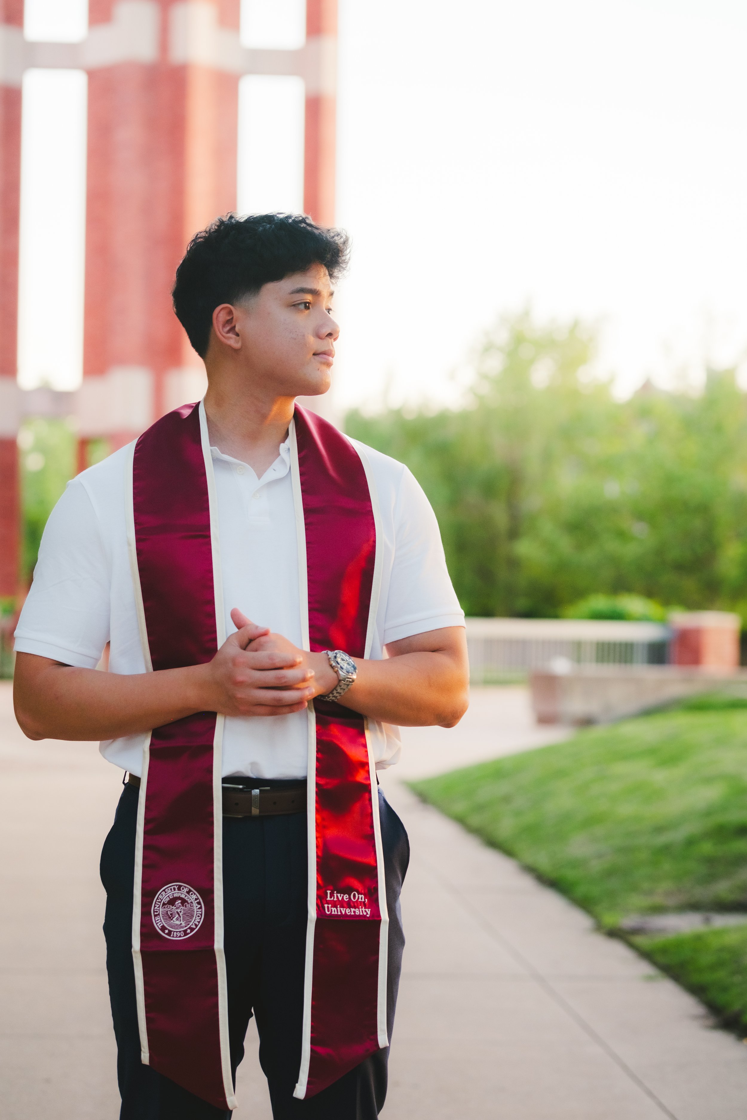 Young man wearing a white polo shirt and a maroon graduation stole standing outdoors on a campus sidewalk, with a red brick building and green trees in the background.
