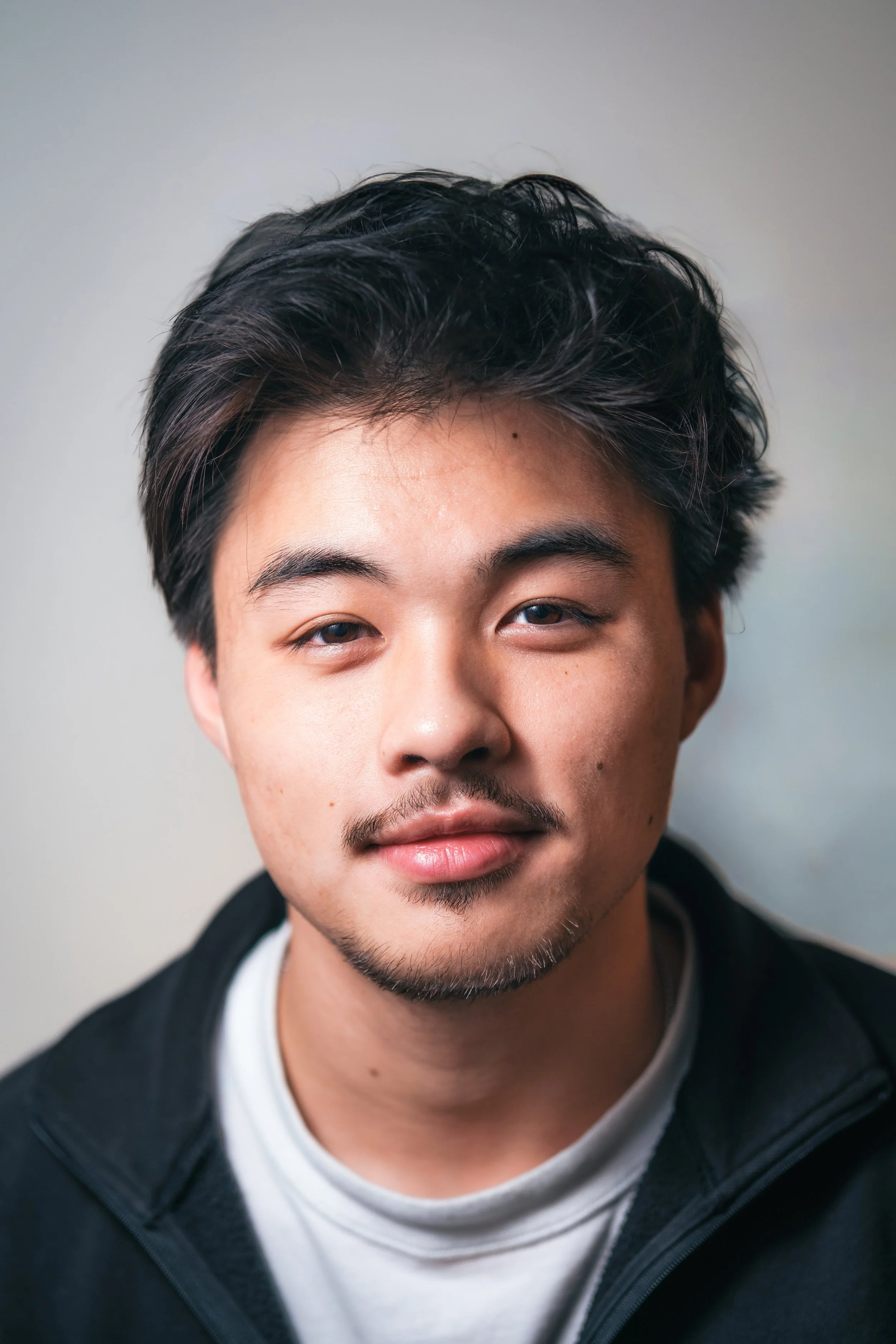 Close-up portrait of a young Asian man with dark, wavy hair, slight facial hair, and a relaxed expression, wearing a black jacket over a white shirt, against a plain out-of-focus background.
