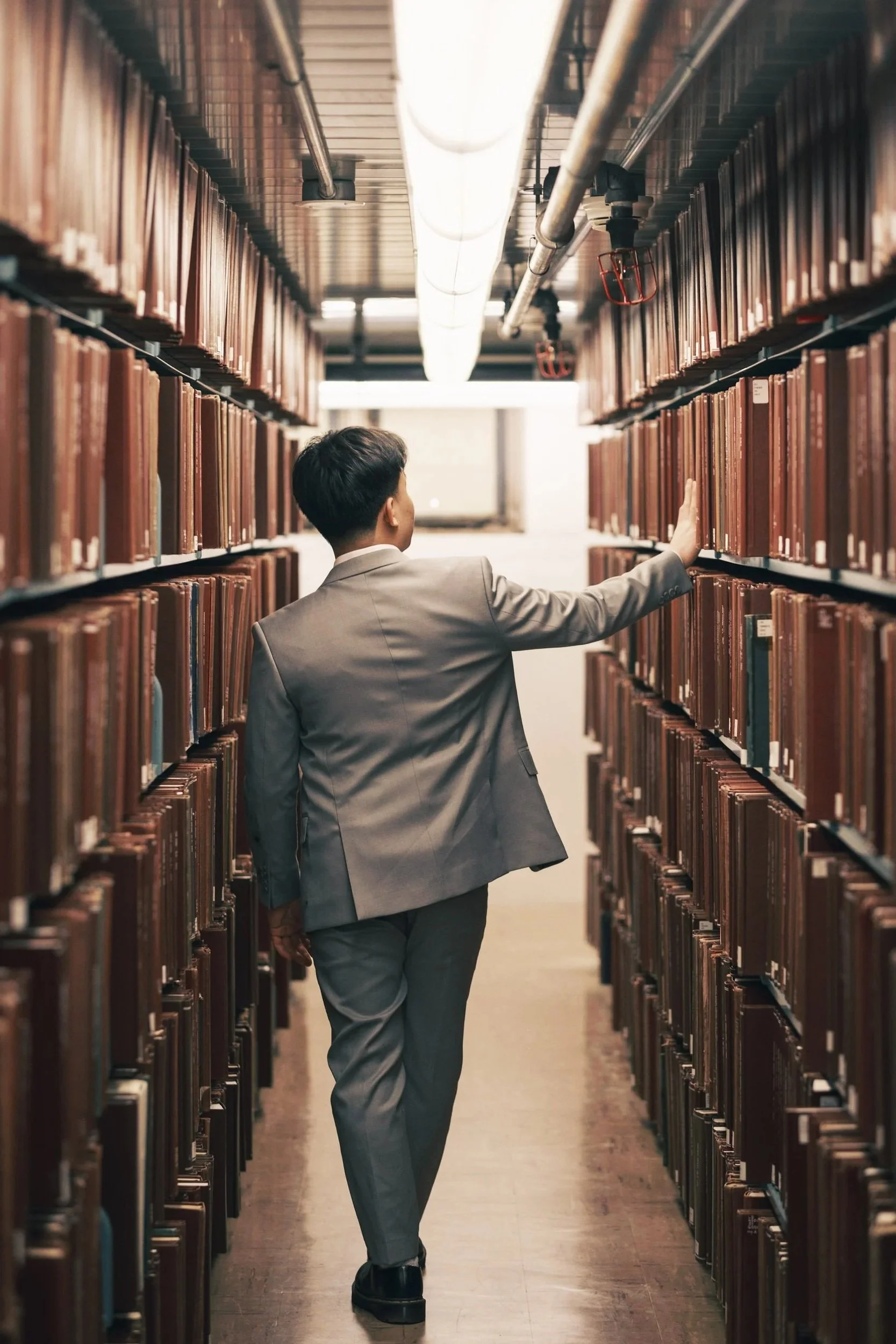 A person in a gray suit walking through a library aisle lined with bookshelves on both sides, reaching out to touch a book on the shelf.
