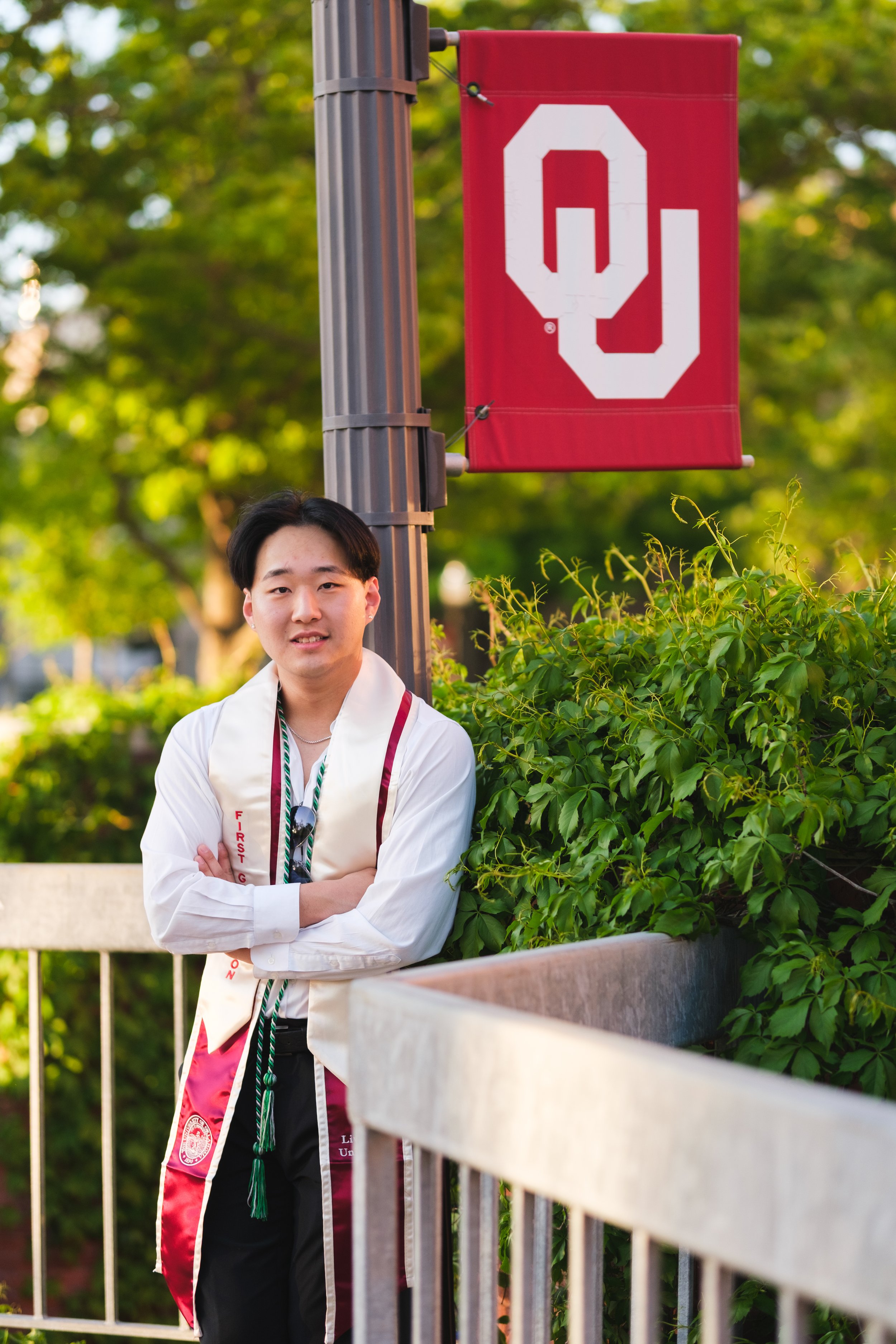 A young man in graduation attire standing outdoors with a red Oklahoma Sooners banner behind him and green foliage around.