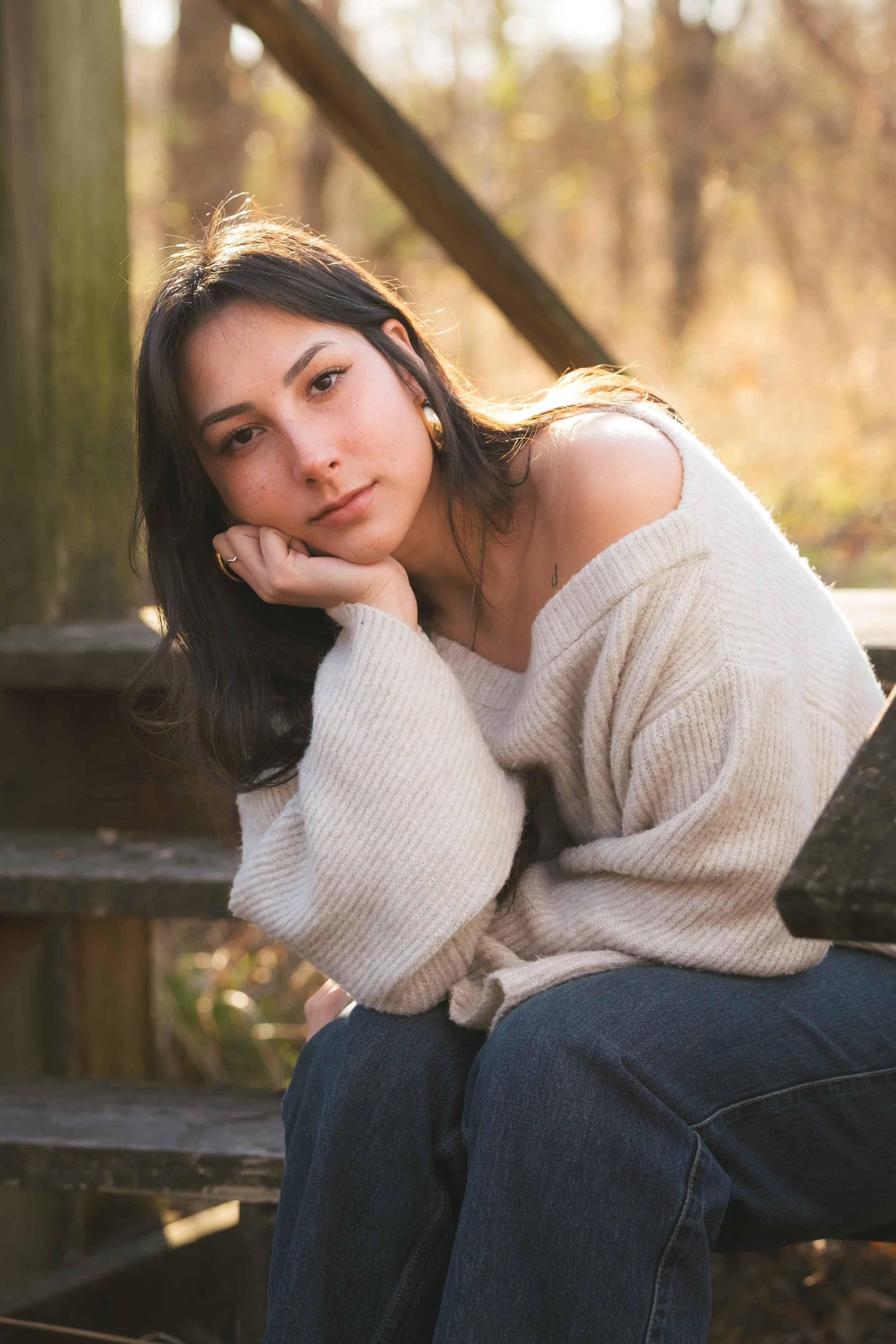 A young woman with dark hair and freckles, wearing a cozy beige sweater and dark jeans, sits on wooden stairs outdoors in a wooded area during autumn, resting her head on her hand and looking at the camera.