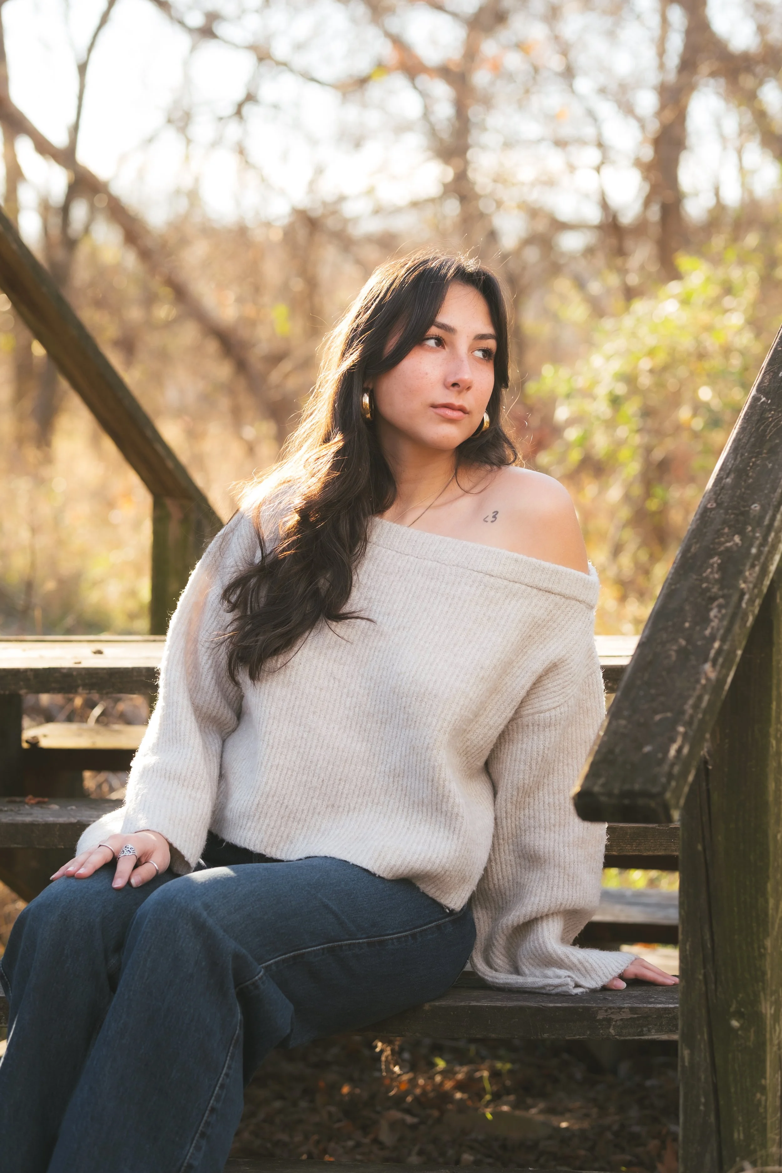 Young woman with dark hair wearing a cream off-shoulder sweater and dark jeans sitting on wooden stairs outdoors during autumn.