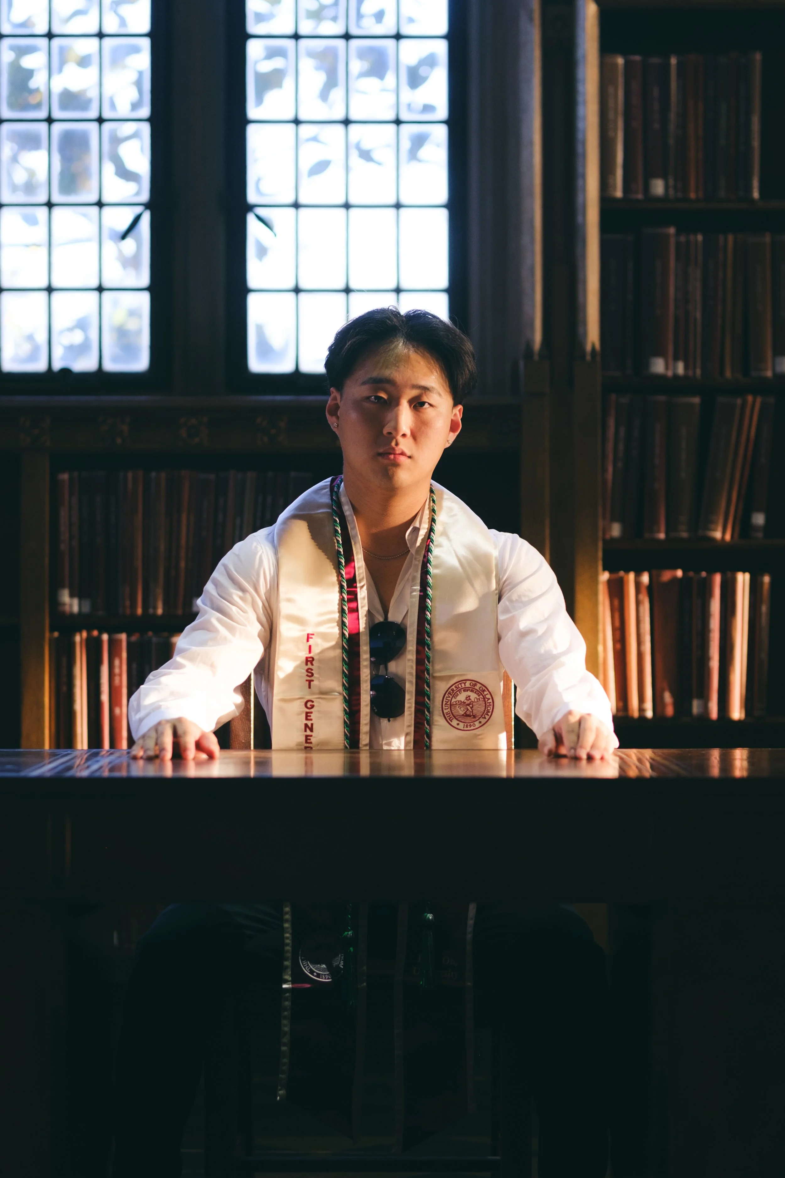 A young man sitting at a wooden table in a library, wearing a white shirt and a graduation stole, with bookshelves and large window behind him.