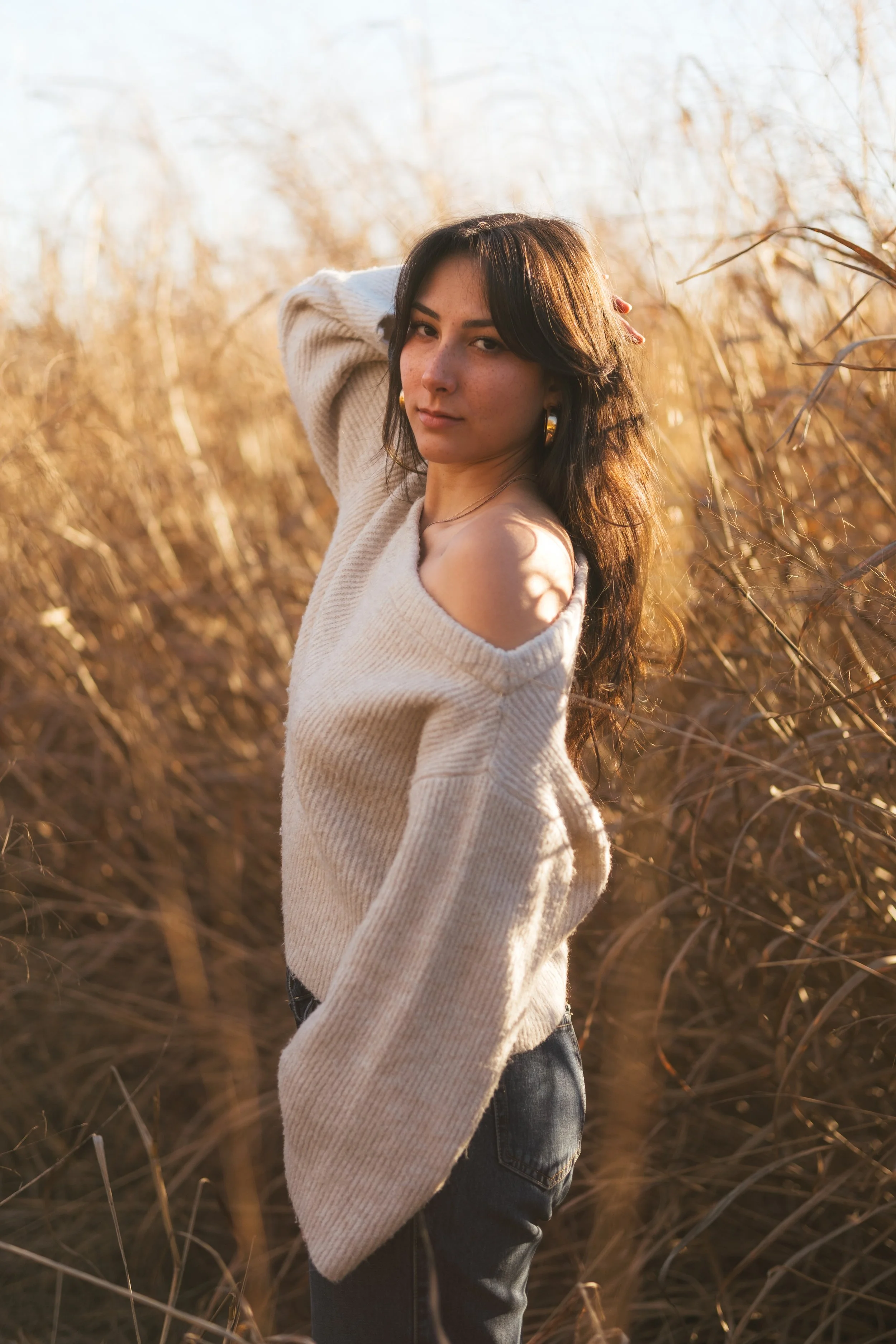 Woman with shoulder-length hair wearing a beige off-shoulder sweater and dark jeans stands in a field of dried tall grass during golden hour.