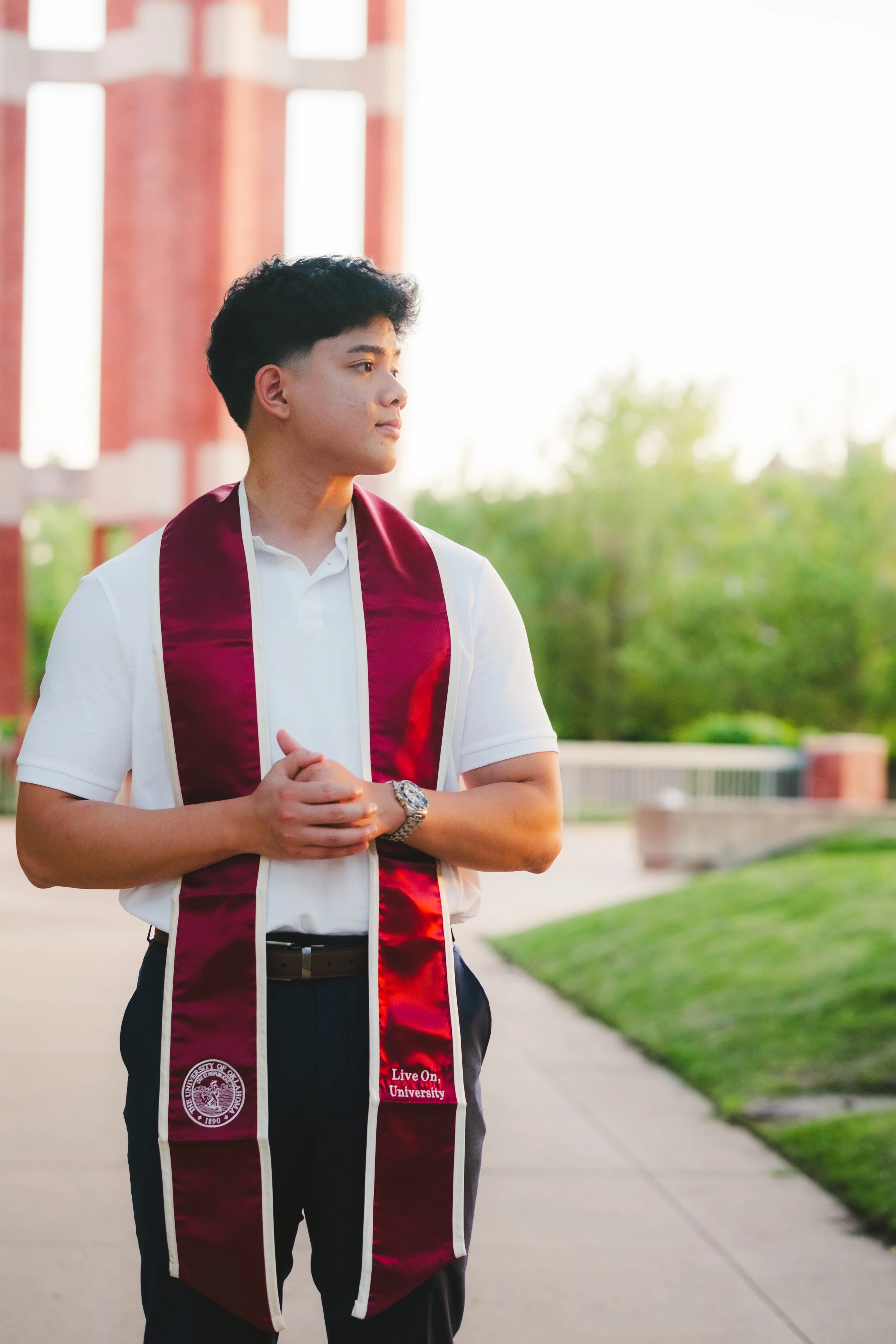 Young man standing outdoors, dressed in a white polo shirt and dark pants, wearing a maroon graduation stole with the University of Arizona emblem, during sunset with greenery and a brick building in the background.
