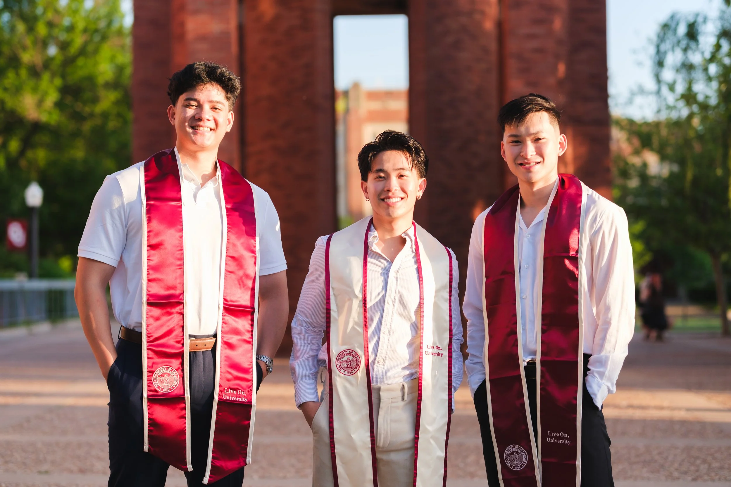 Three young men standing outdoors, smiling, wearing graduation sashes, with a brick arch and green trees in the background.