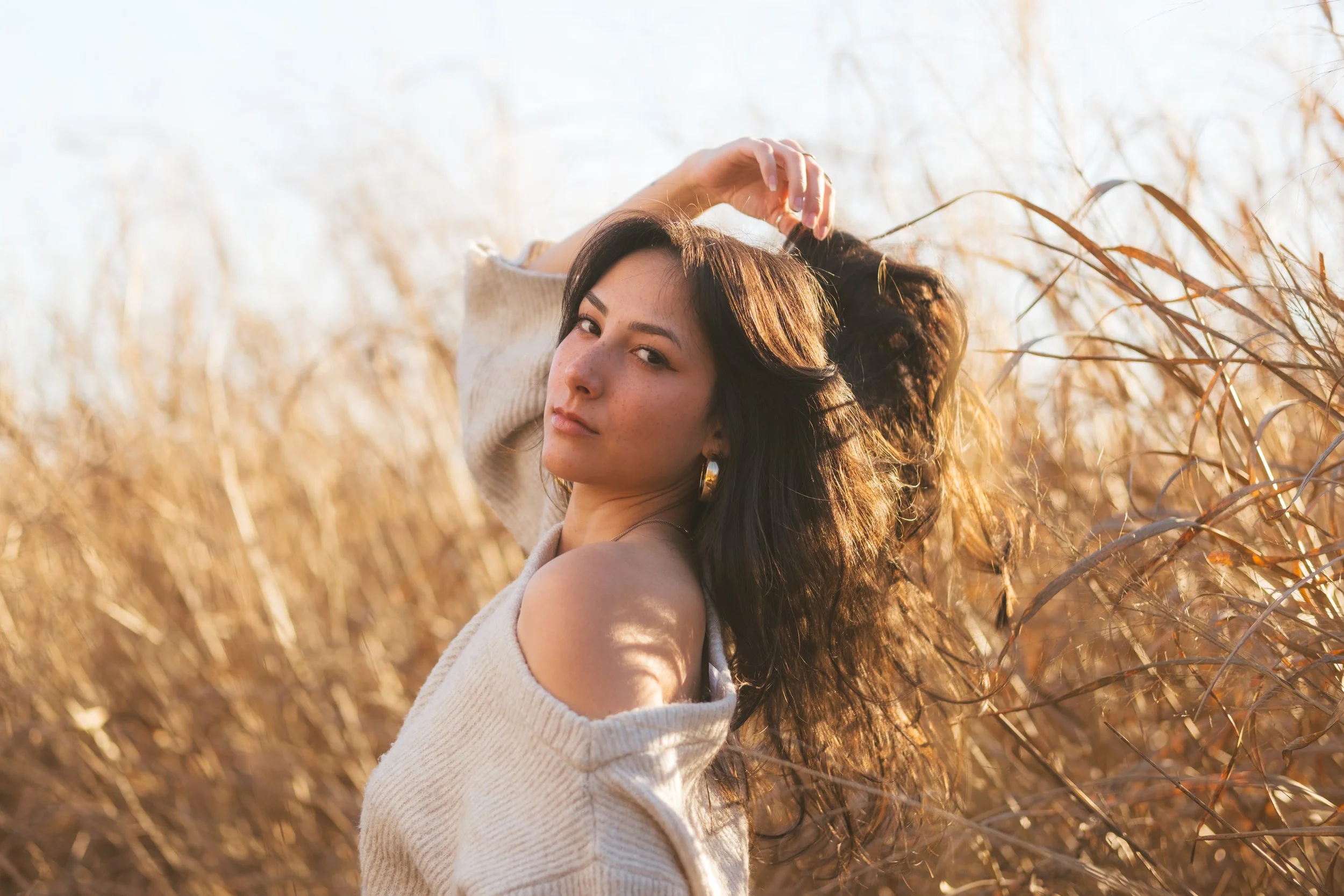 A woman standing in a field of tall, dry grass during golden hour, with one arm raised over her head and looking directly at the camera.