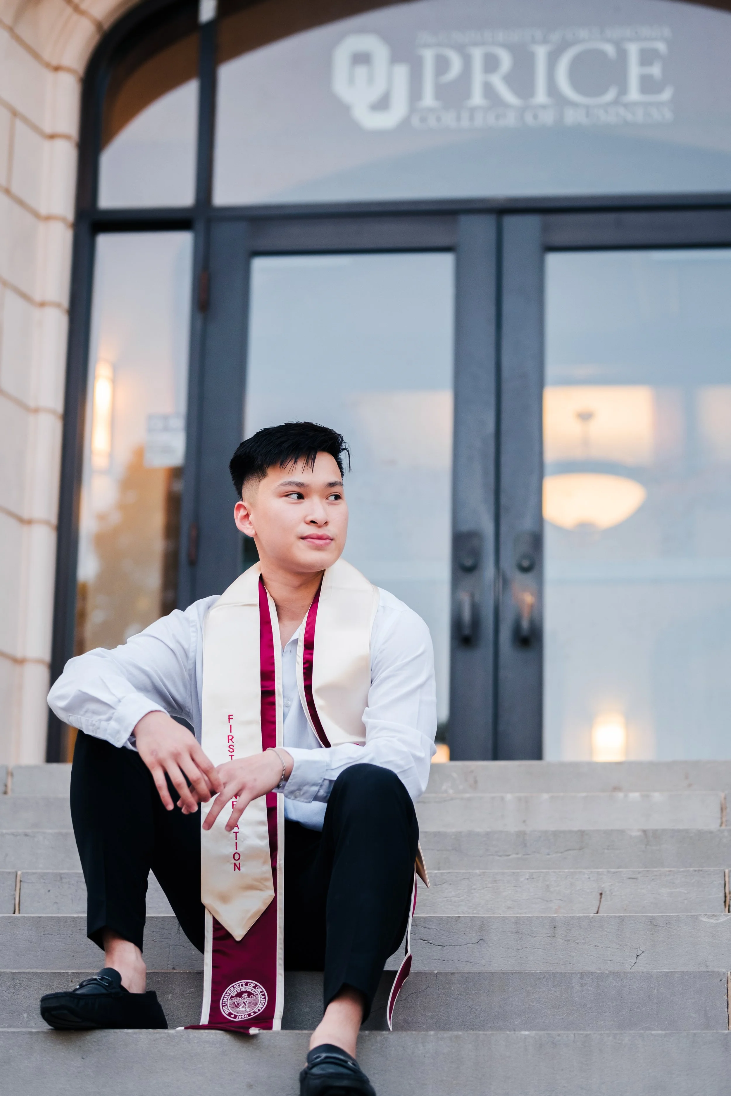 A young man sitting on steps outside a building with a University of Oklahoma Price College of Business sign, wearing a white shirt, black pants, and a cream-colored graduation stole with maroon accents.