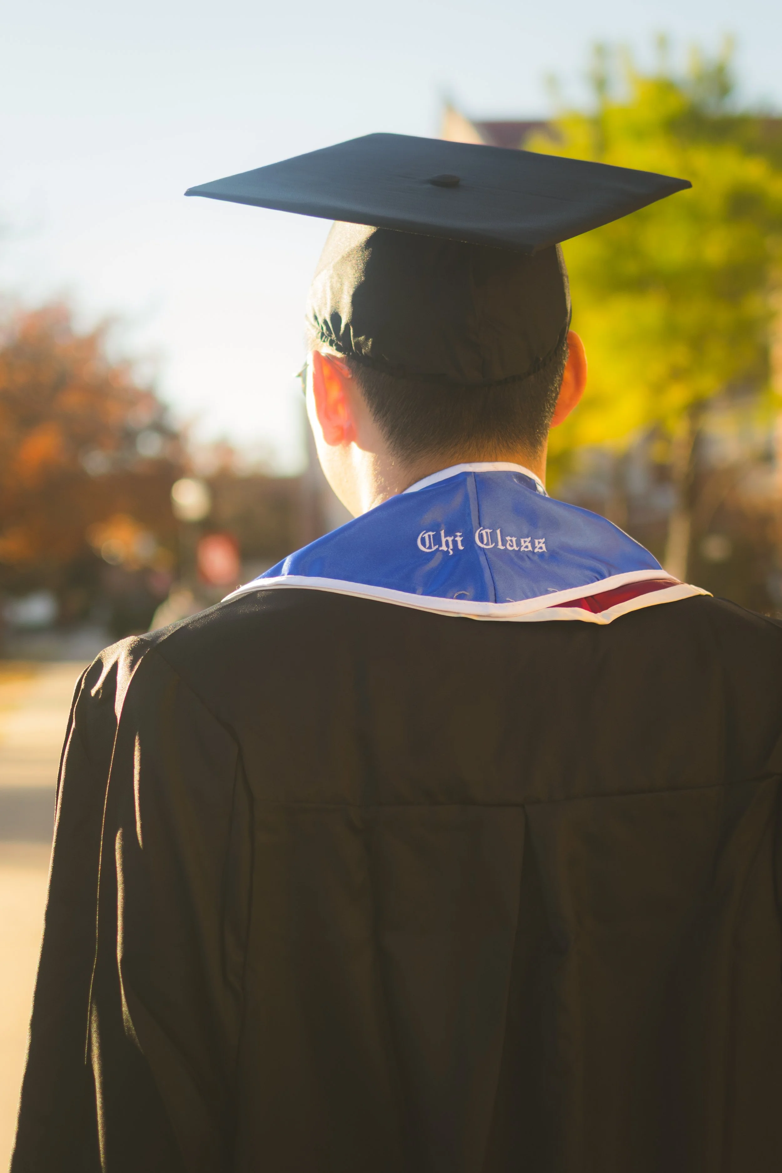 Back view of a graduate wearing a black cap and gown with a blue stole that says "The Class" and a white collar, outdoors in sunlight with trees in the background.