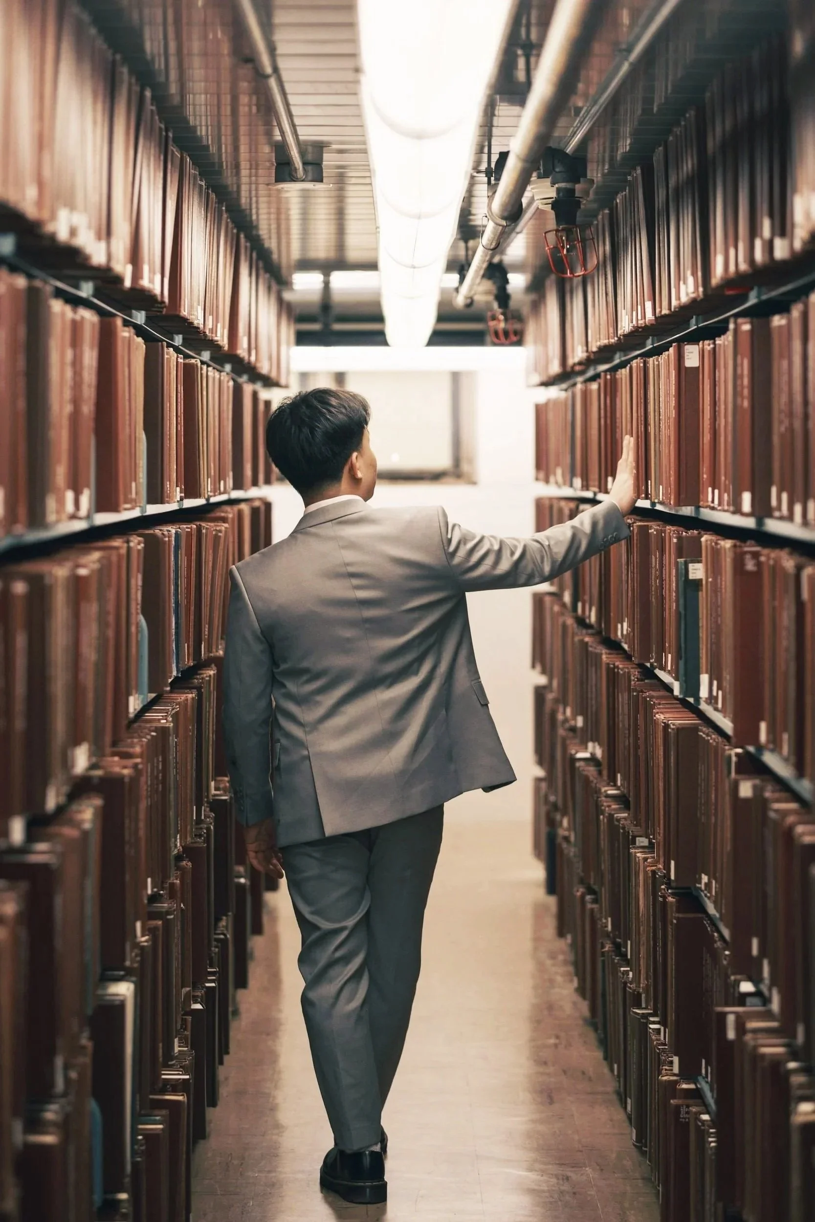 A man in a gray suit walking through a library aisle, reaching out to touch books on the shelves.
