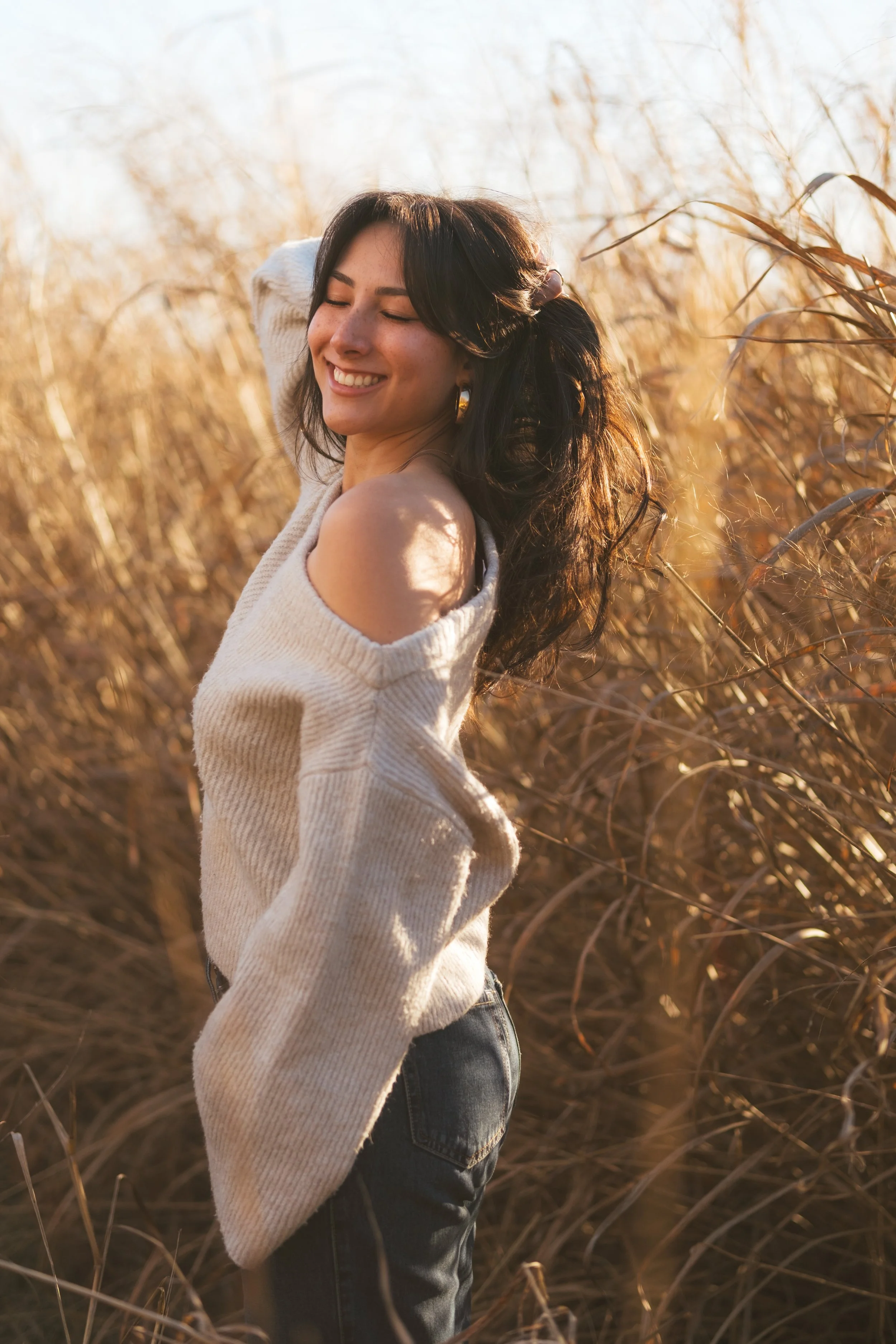 A woman with dark hair posing in a field of tall, dry grass during golden hour, smiling with her eyes closed, wearing a cream off-shoulder sweater and dark jeans.