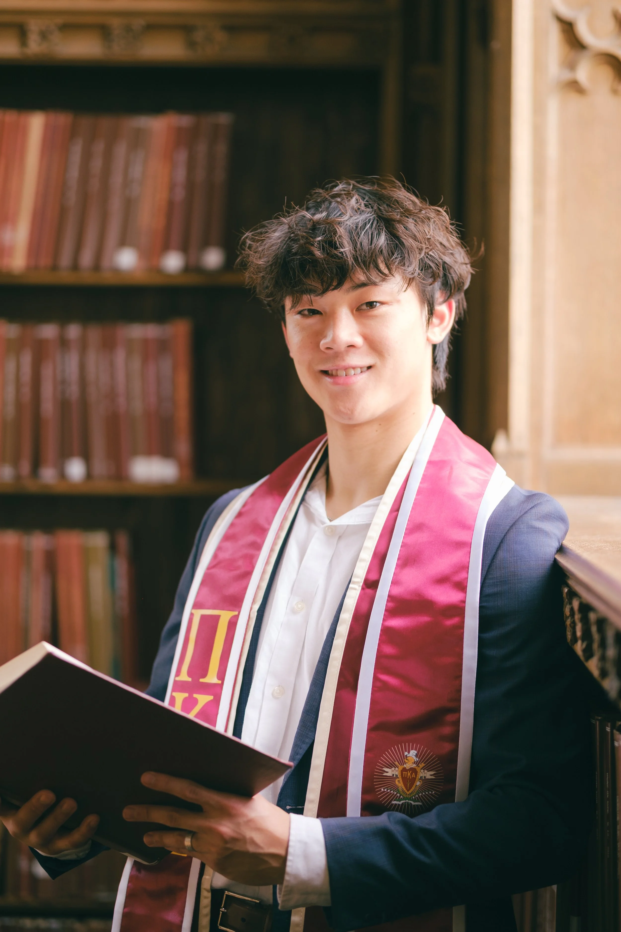 A young man in formal attire holding a diploma or certificate, wearing a maroon and gold stole, standing in a library with wooden shelves of books.