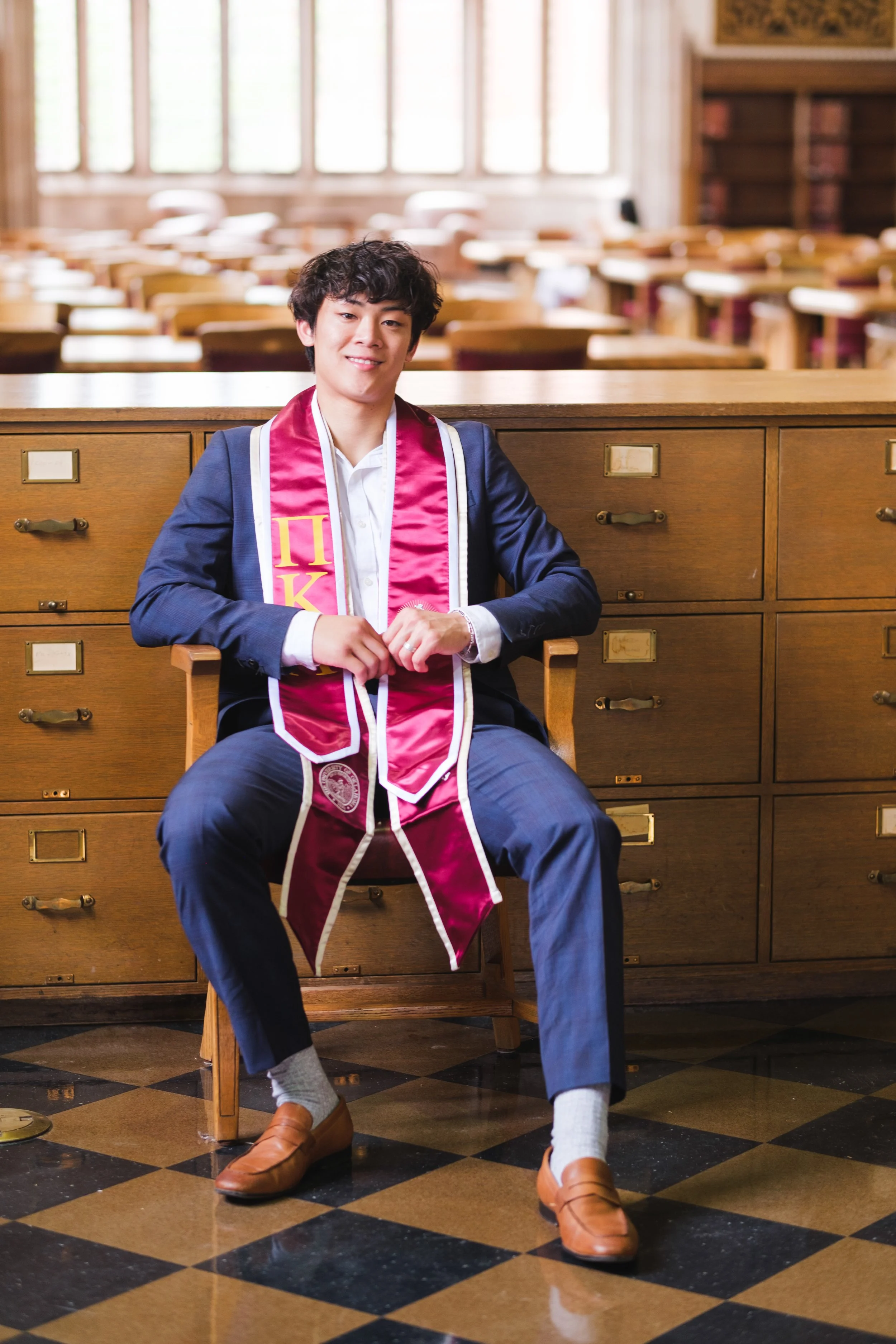 A young man in a graduation gown and stole sitting in an office with a large wooden cabinet behind him, in a bright room with large windows, smiling at the camera.