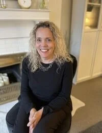 A woman with curly blonde hair, wearing a black top and a necklace, smiling while sitting on a chair in a cozy, well-lit room.