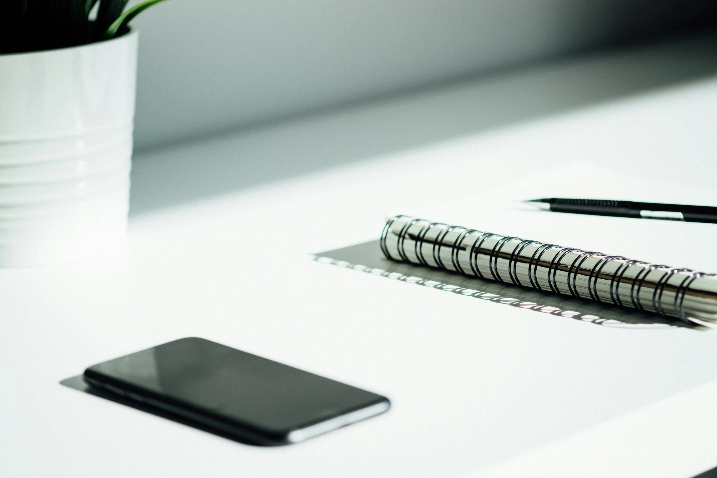 Minimalist workspace with a smartphone, a black pen, a spiral notebook, and a small potted plant on a white desk with sunlight casting shadows.