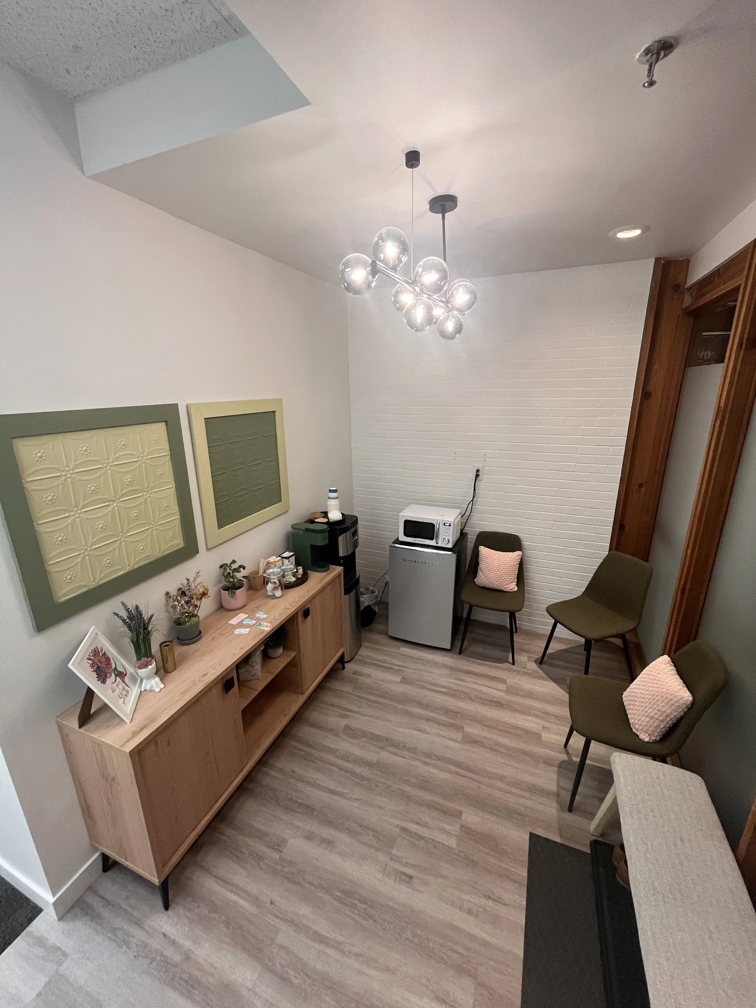 An indoor waiting area with three olive-green chairs with pink pillows, a wooden sideboard with plants and framed artwork, a coffee machine, a microwave, a mini-fridge, and a modern chandelier with multiple glass globes.