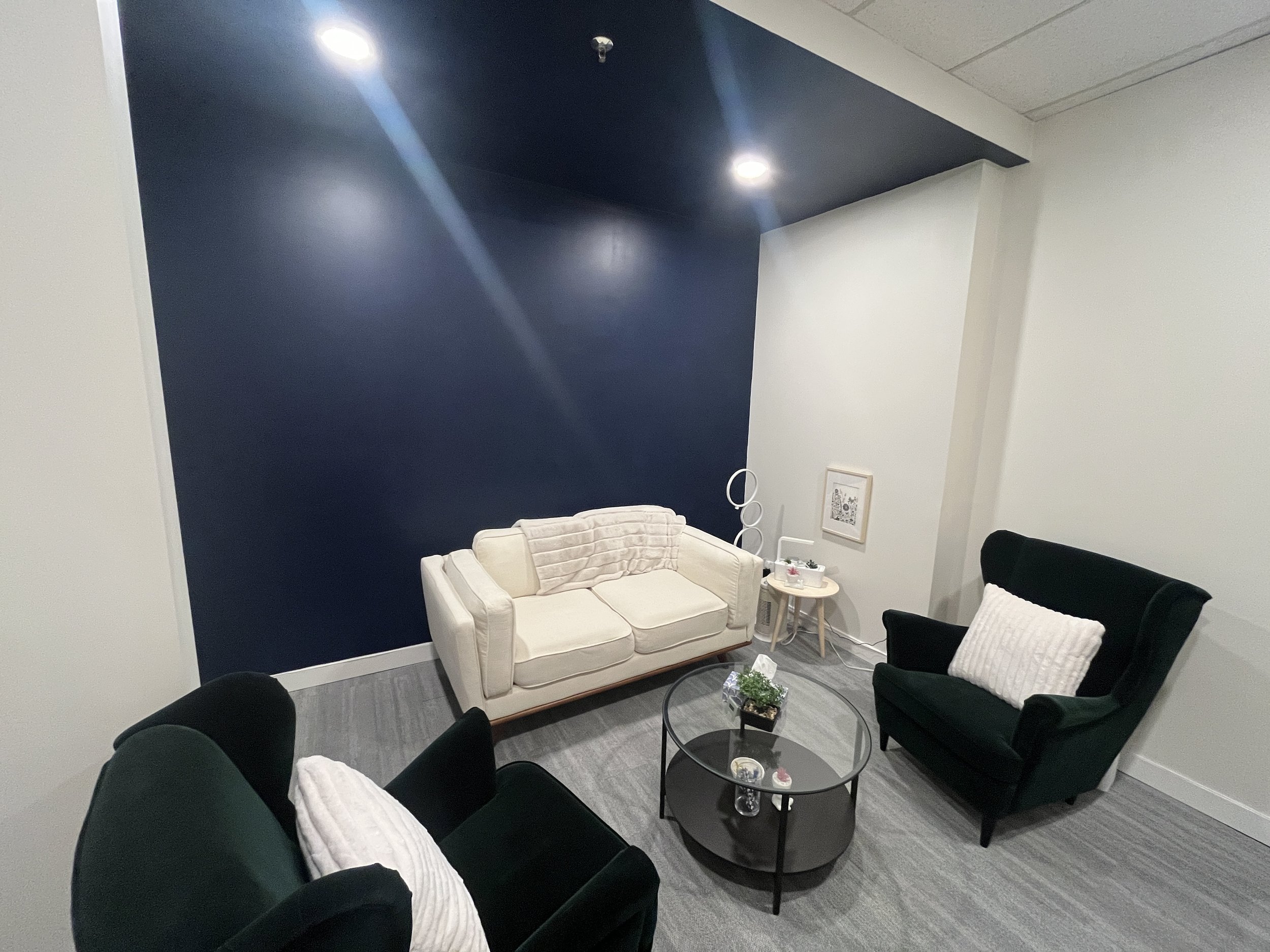 A cozy living room corner with a white sofa, and two black armchairs with white pillows, a round glass coffee table with decorative items, a small side table, and framed artwork on the wall, with blue accent wall and ceiling lights.