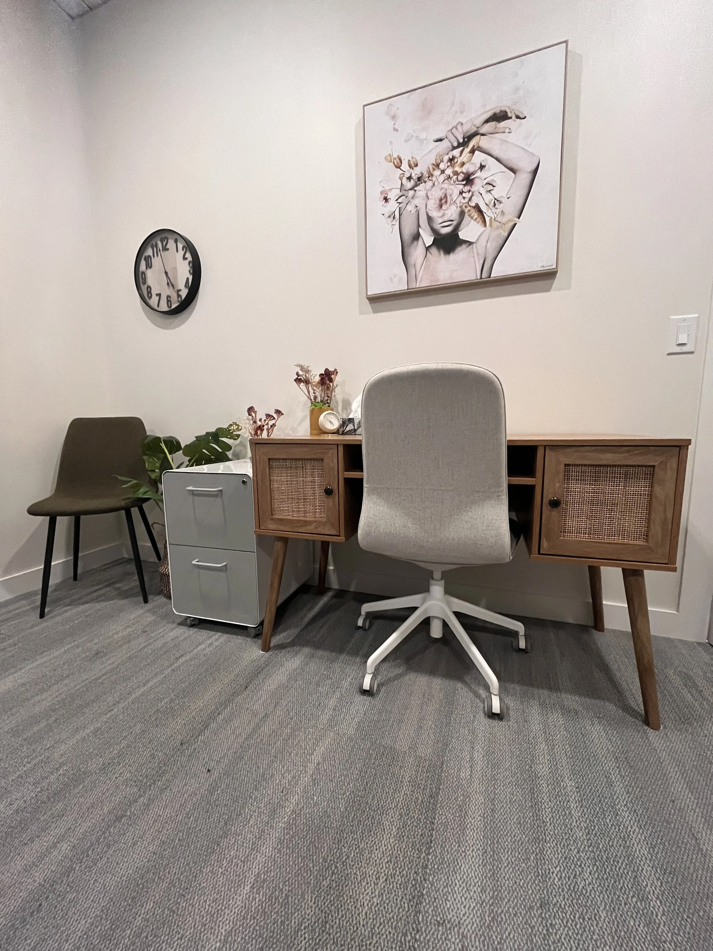An office corner with a wooden desk, an office chair, a brown accent chair, a white file cabinet, a potted plant, a vase with dried flowers, a small clock, a large wall art of a woman with flowers covering her eyes, and a wall switch.