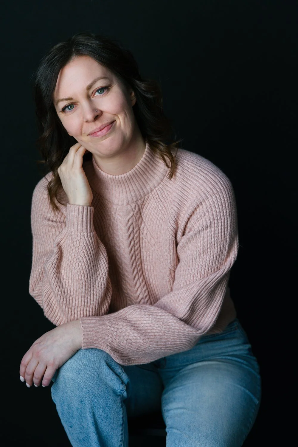 A woman with shoulder-length dark hair wearing a pink sweater and blue jeans, sitting against a black background, smiling softly at the camera.