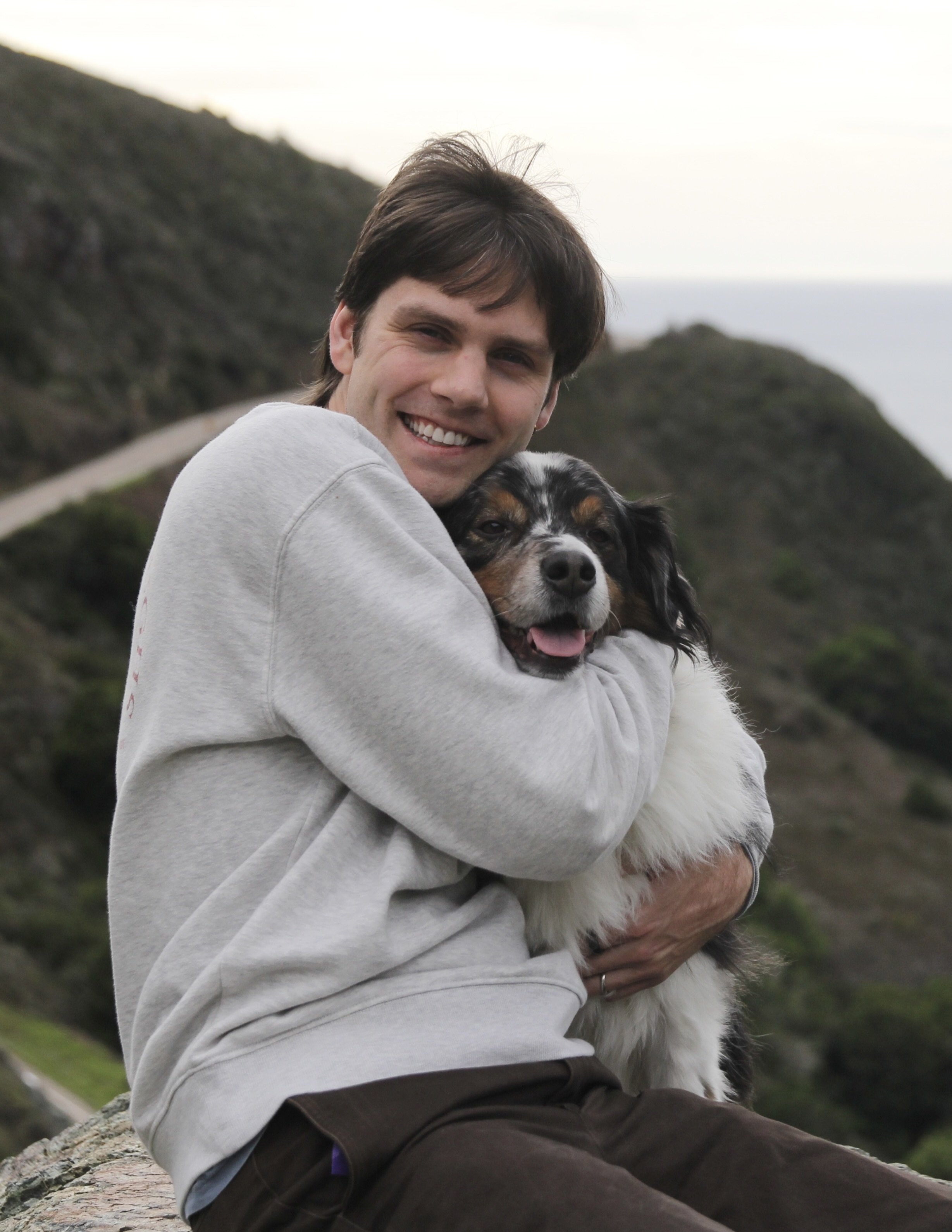 A man with dark hair smiling and holding a black, white, and brown dog while sitting outdoors on a rocky surface, with green hills and cloudy sky in the background.