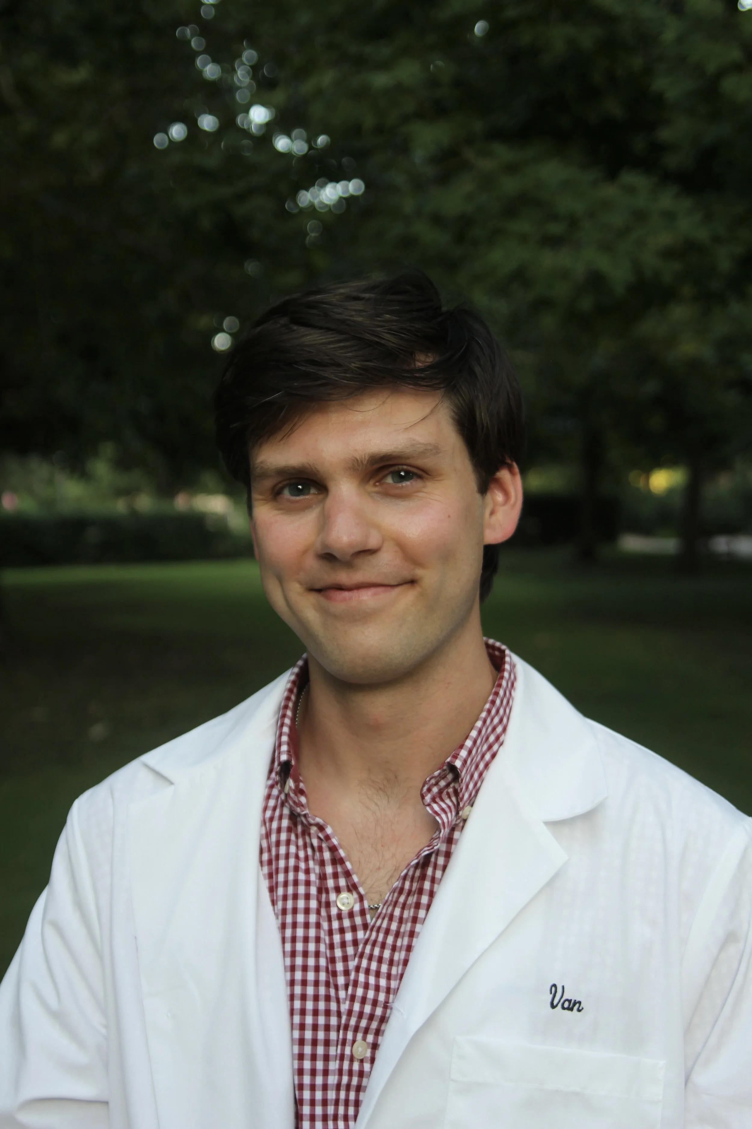 Young man with dark hair in a white coat and red checkered shirt standing outdoors in a park with trees background.