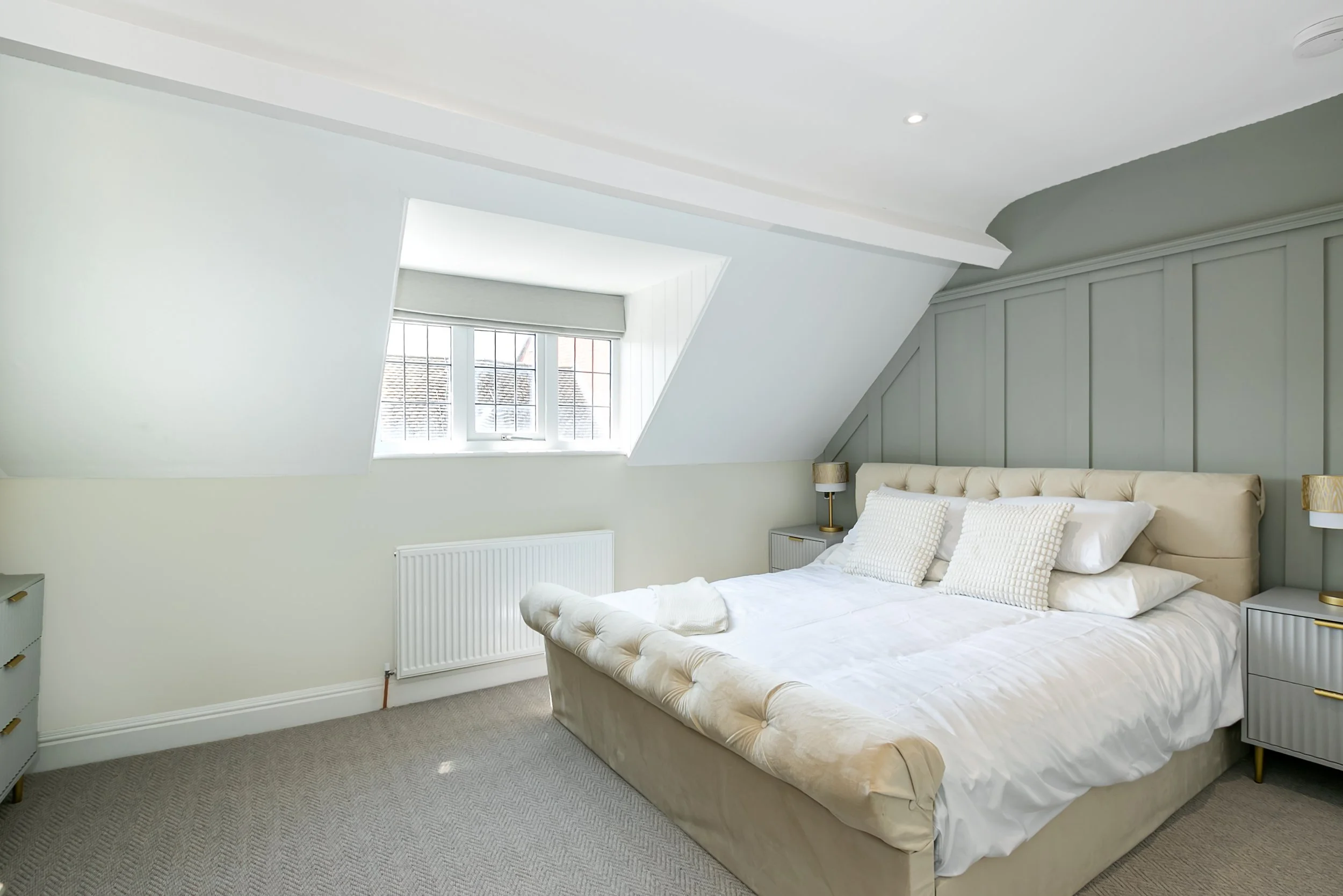 A bedroom with a beige upholstered bed, white bedding, and pillows, flanked by two gray nightstands with gold accents, and a skylight window with blinds on the slanted ceiling.