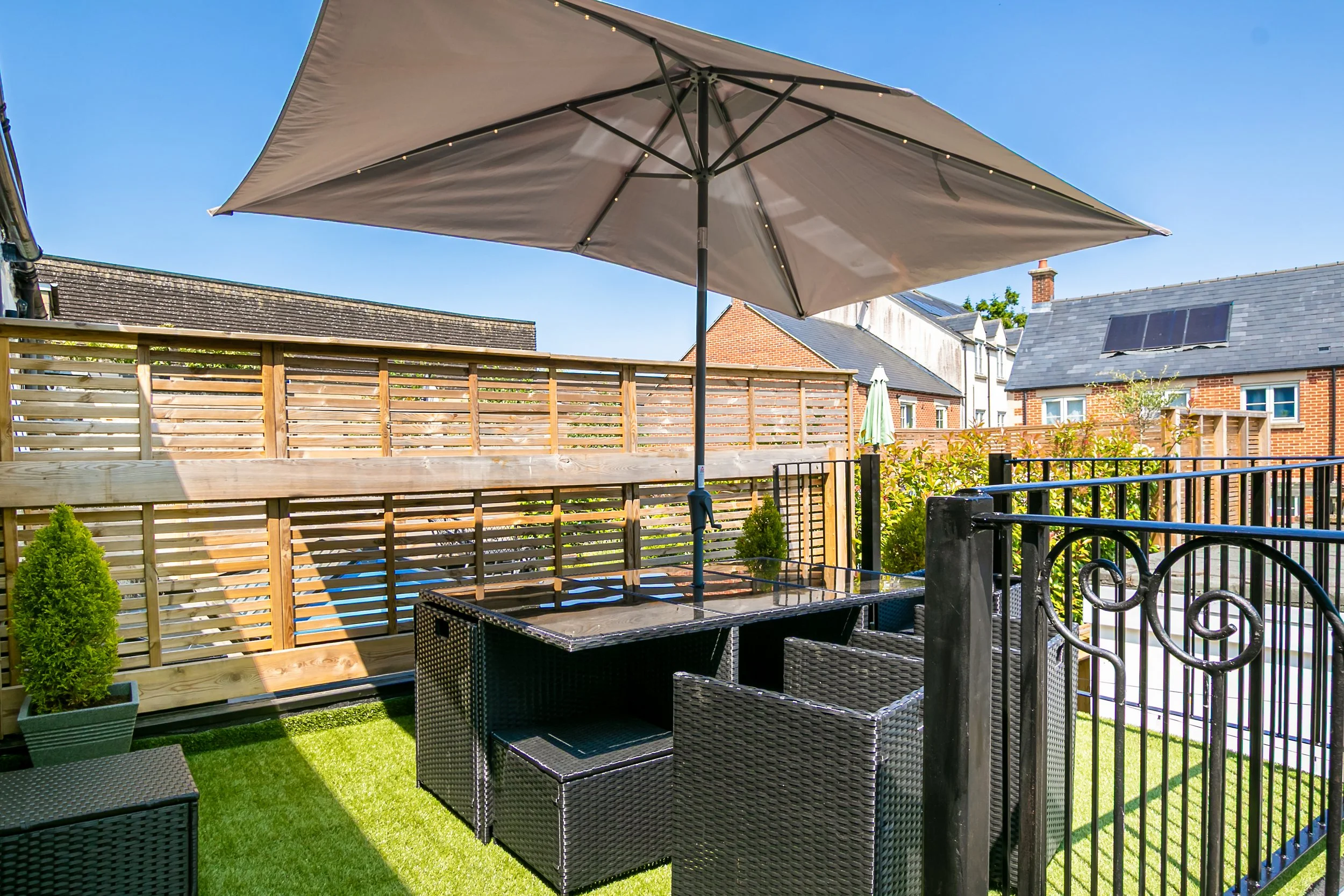 Outdoor patio with a large beige umbrella, black wicker table and chairs, small potted bushes, and wooden fence, surrounded by houses under a clear blue sky.
