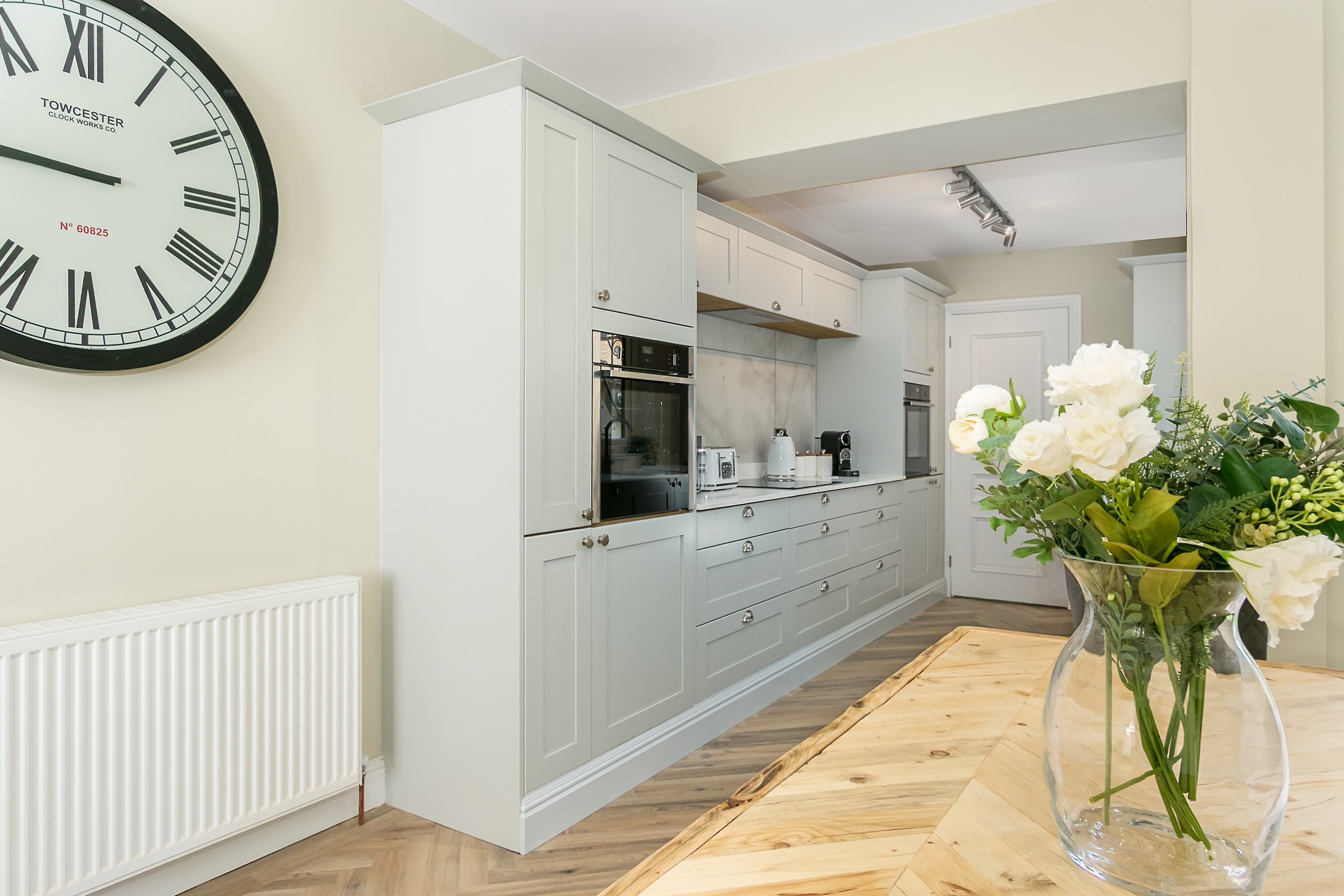 A modern kitchen with white cabinetry, a large wall clock, and a wooden dining table with a glass vase of white flowers.