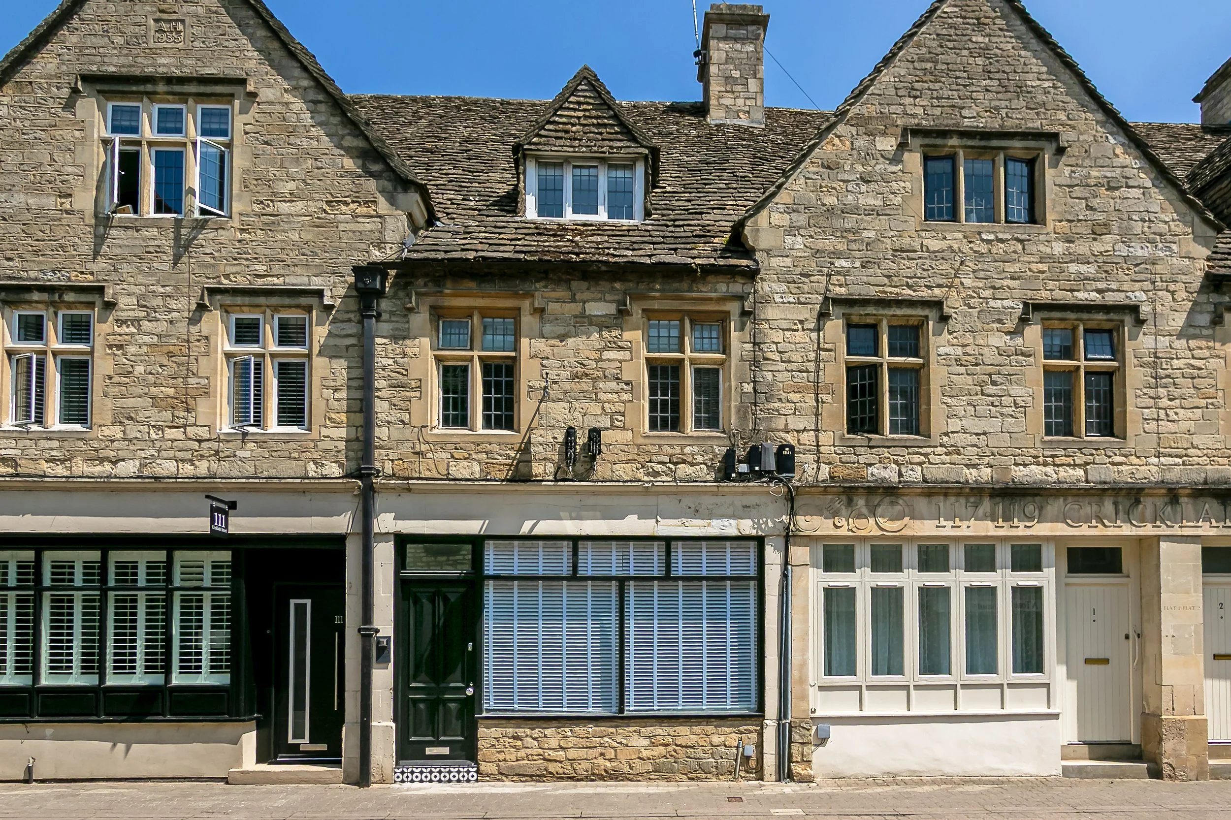 Street view of a historic stone building with several windows, some with shutters, and ground-level shops with black and green doors and window frames.