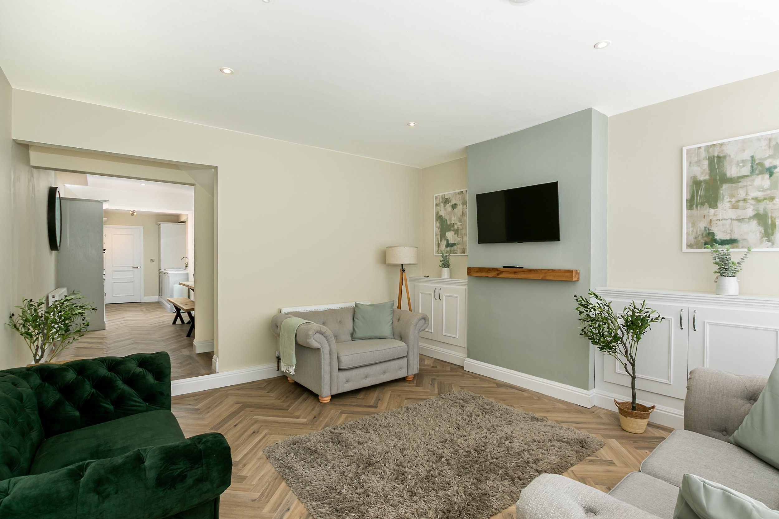 Living room with beige walls, a flat-screen TV mounted on a light green accent wall, white cabinetry, a beige couch, a dark green velvet armchair, potted plants, abstract artwork, a wooden floating shelf, a rug, and a floor lamp.