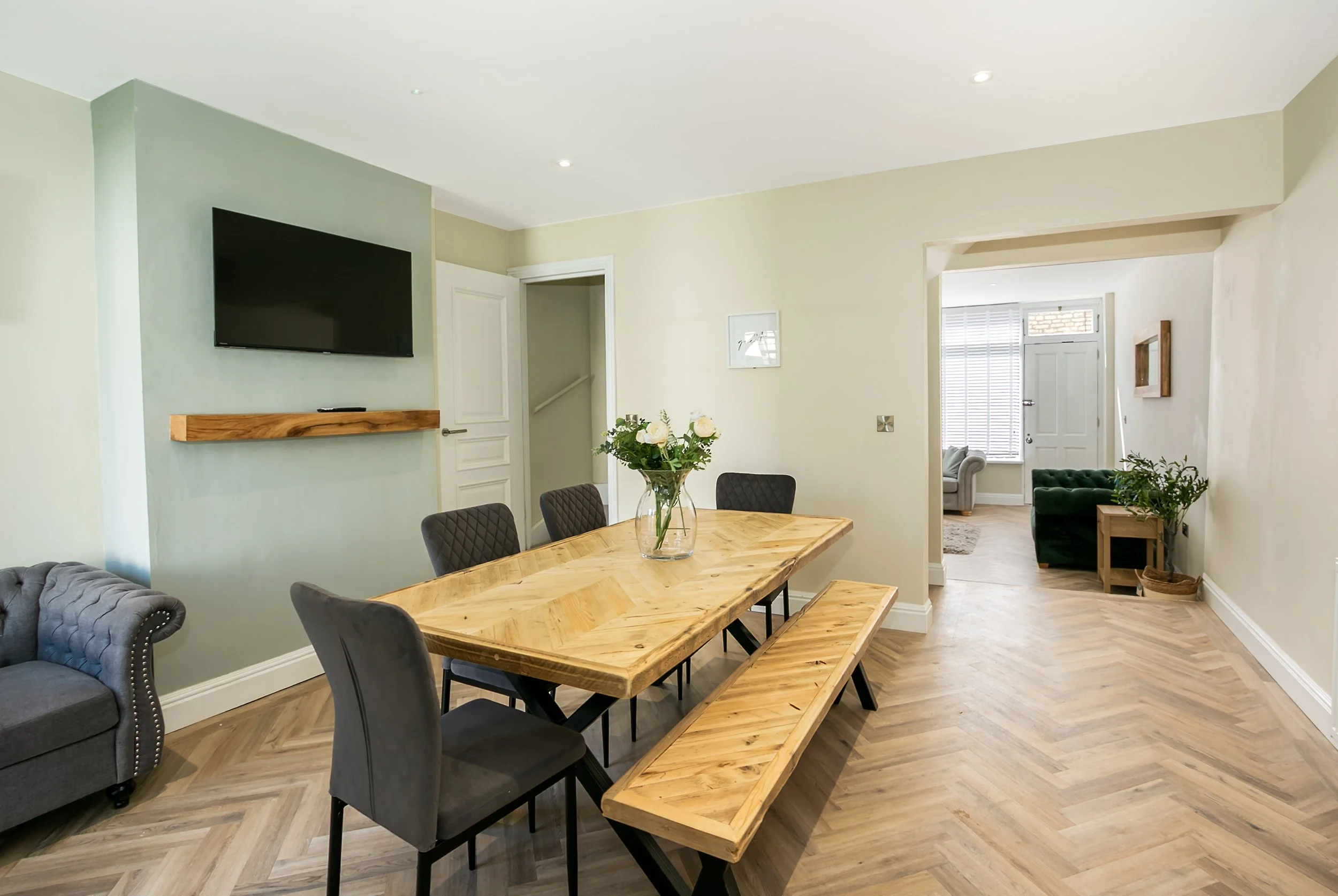 A dining room with a wooden table, six chairs, a vase with white roses, a wall-mounted TV, and an opening leading to a living area with a green armchair and window with blinds.