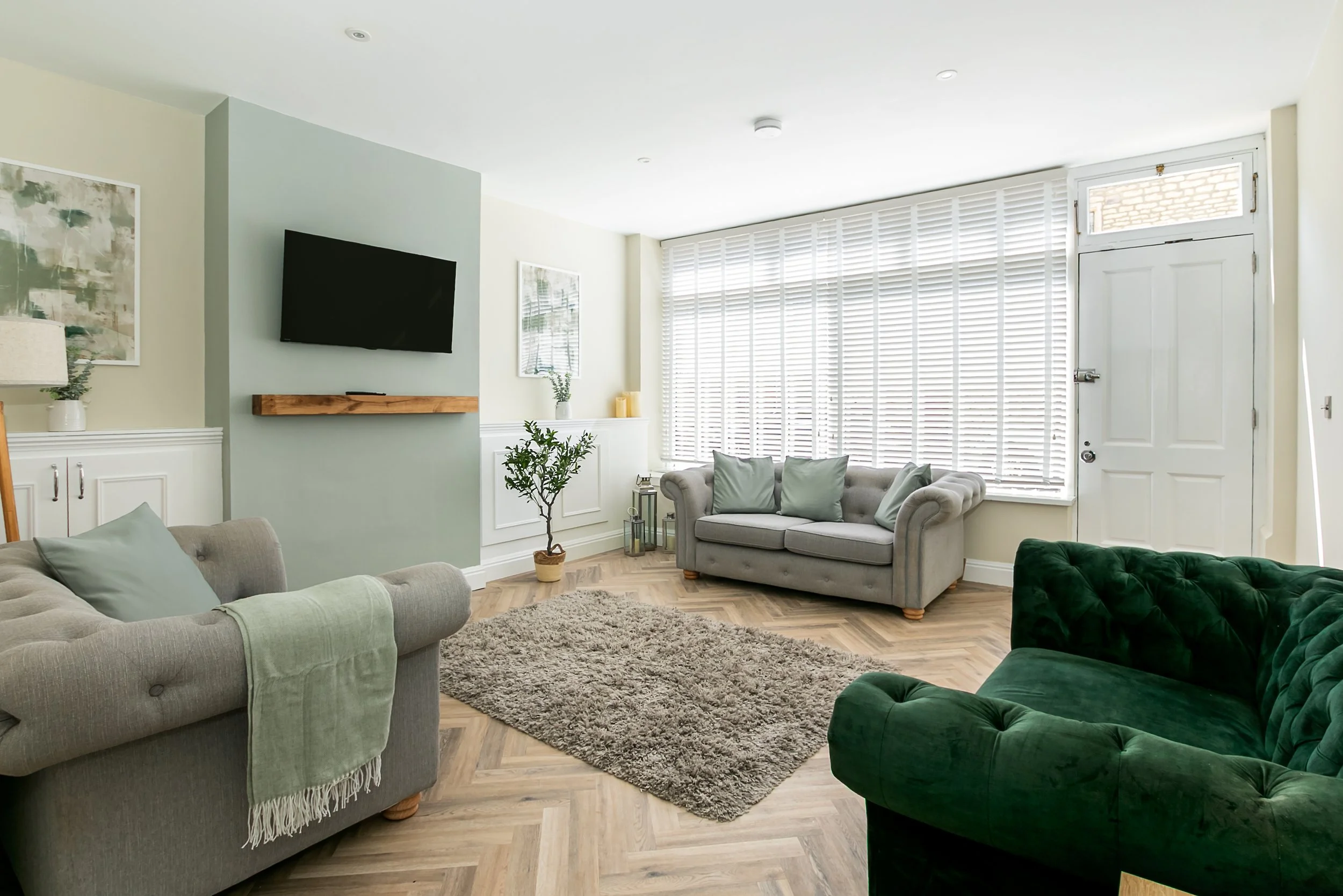 Living room with gray and green sofas, a shaggy rug, potted plant, wall-mounted TV, large window with blinds, and white door.