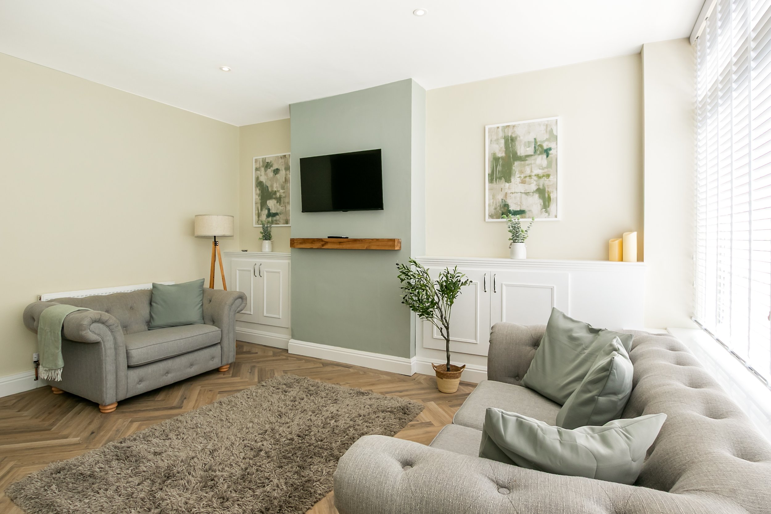 Living room with two gray sofas, a rug, a potted plant, wall art, a wall-mounted TV, and built-in cabinets with decorative items, natural light from window blinds.