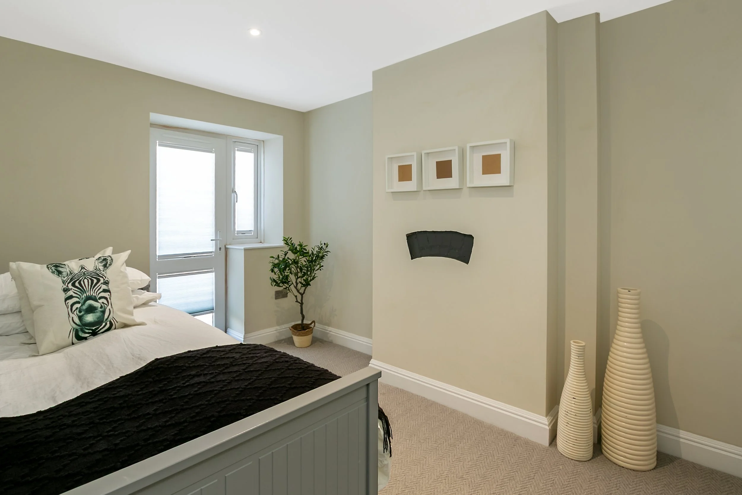 A bedroom with a bed, white bedding, zebra pillow, potted plant, window, framed artwork, decorative vases, and wall-mounted fireplace.
