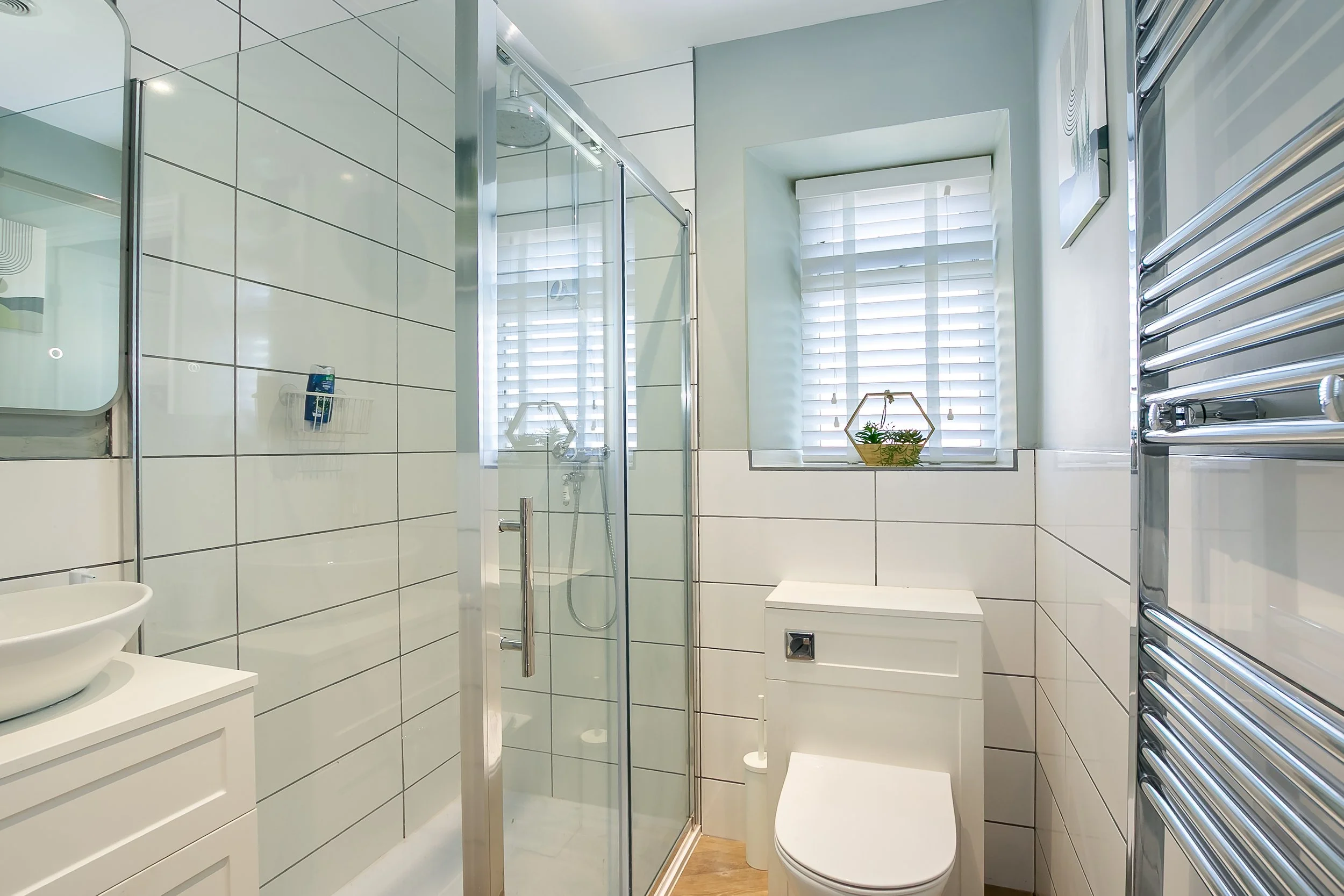 A modern bathroom with white tiled walls, a glass-enclosed shower, a small window with white blinds, a white toilet, a wall-mounted towel radiator, and a small potted plant on the windowsill.
