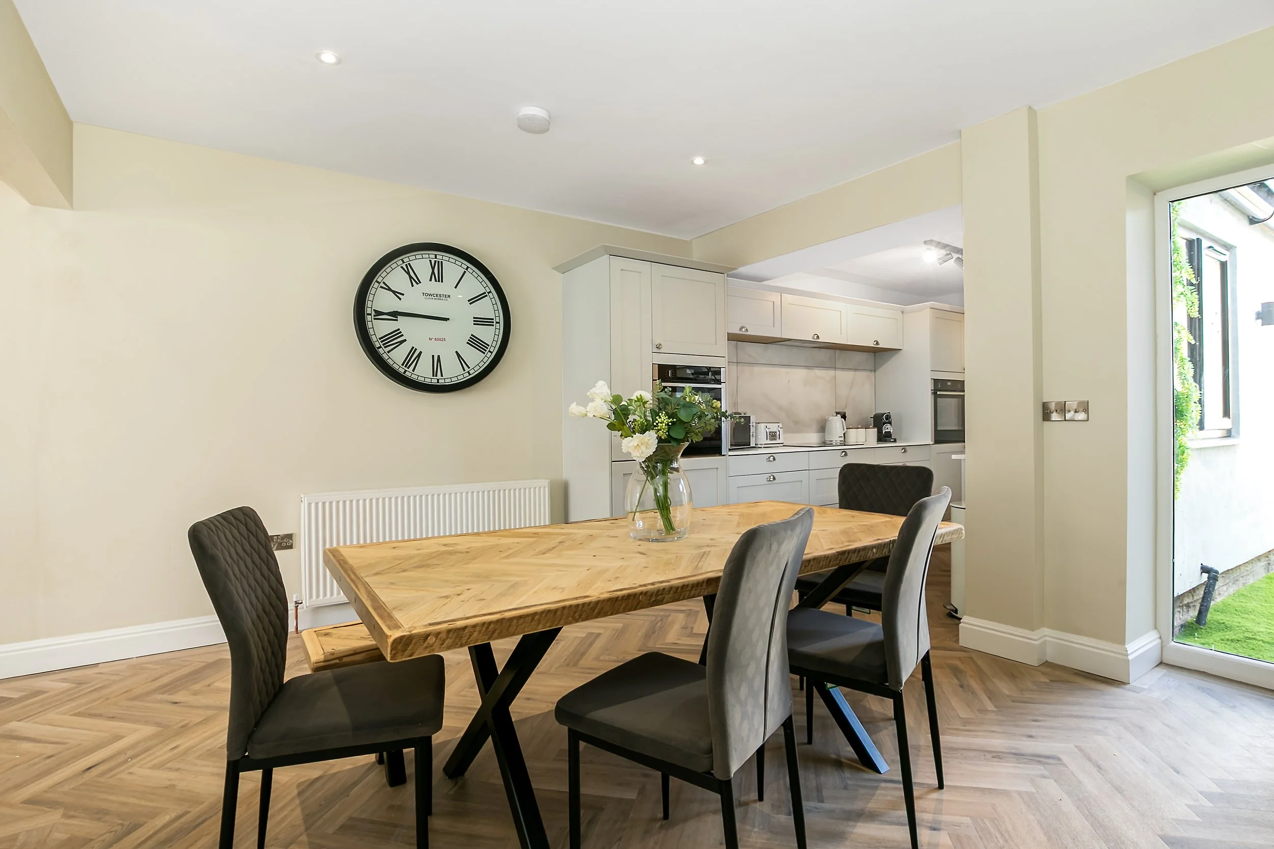 Modern dining room with a wooden table, four chairs, a large wall clock, and an open kitchen with white cabinets and appliances.