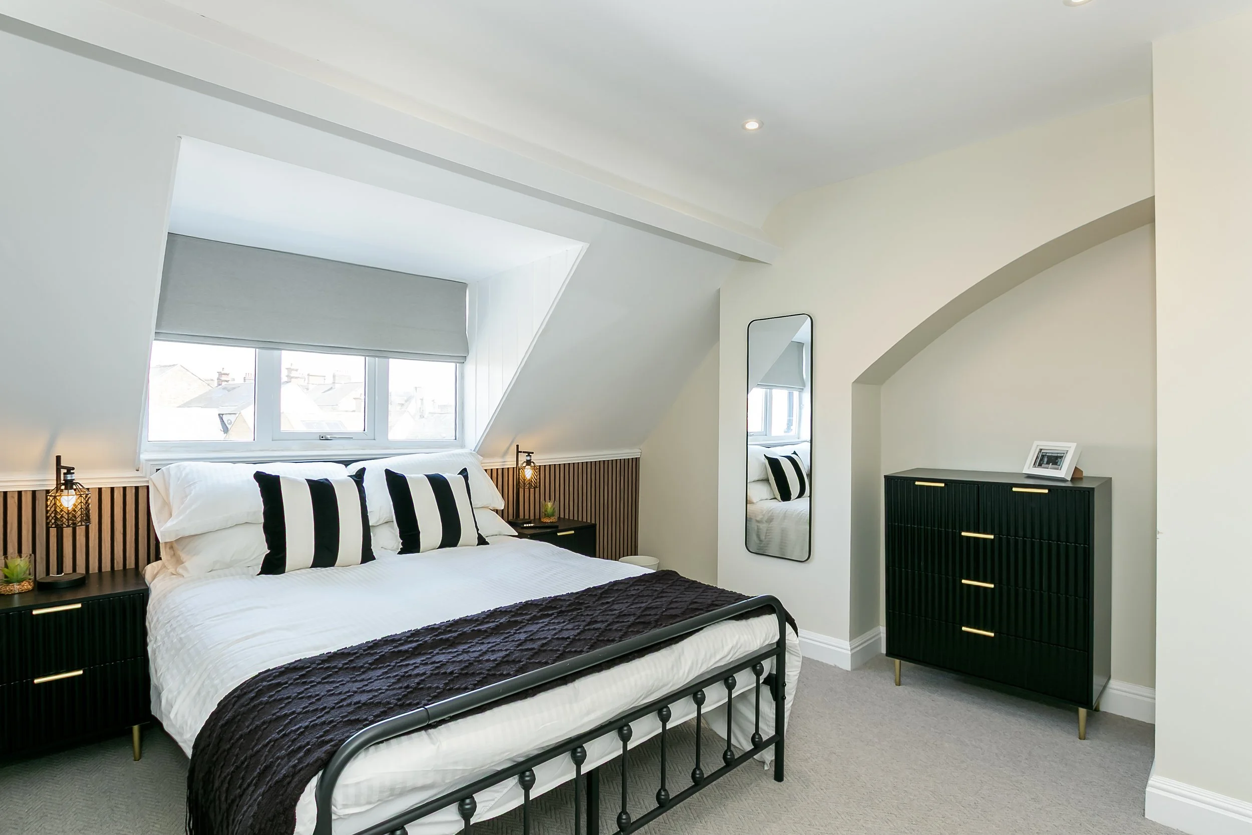 A modern bedroom with a black metal bed frame, white bedding, and black and white striped pillows. Two black nightstands with gold accents and table lamps flank the bed. A large window with a gray blind is above the bed, and a tall mirror and black d