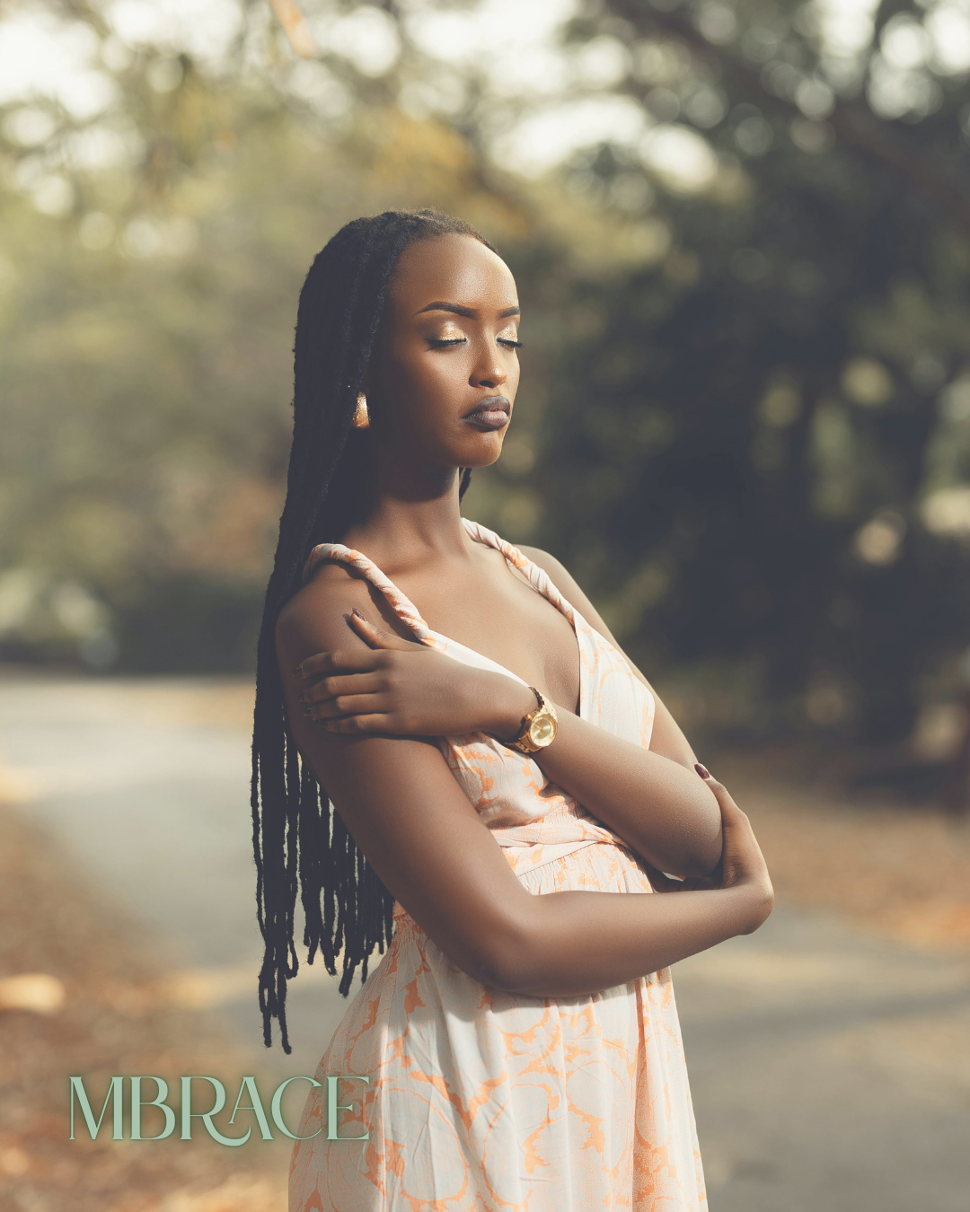 A young woman with dreadlocks standing outdoors with her eyes closed and arms crossed, wearing a floral dress and a gold watch, with trees in the background.