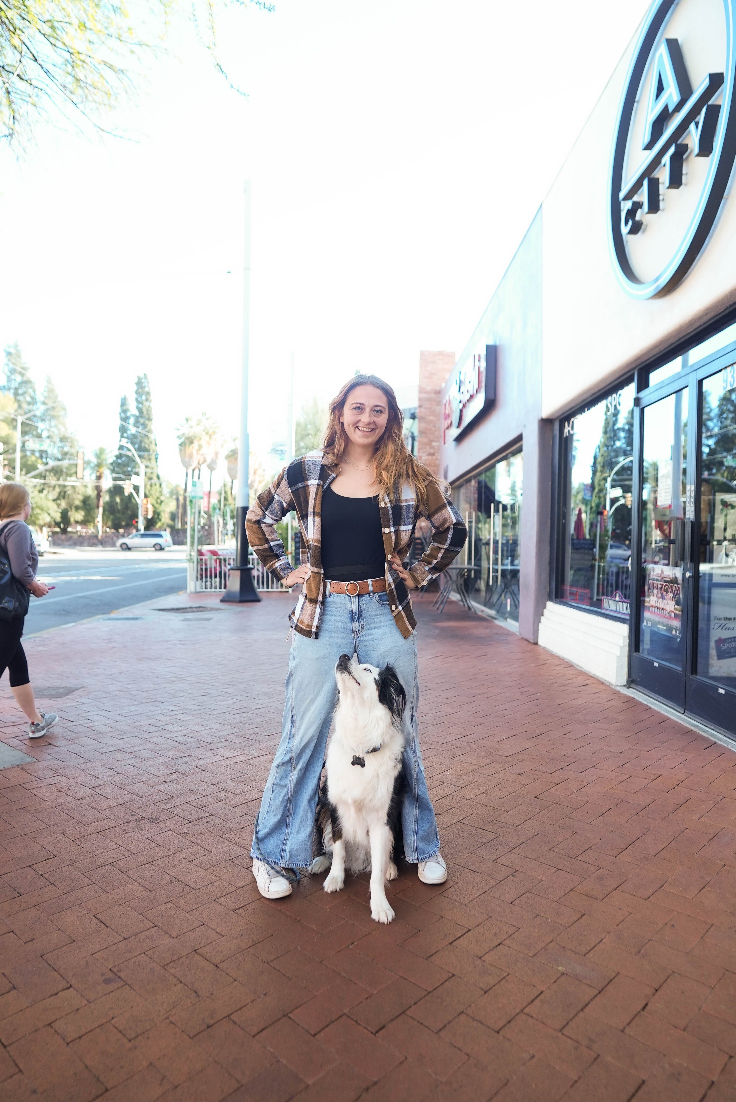 A young woman standing on a sidewalk with her border collie dog in front of an Auto Everything store, smiling at the camera, dressed in a plaid shirt and jeans, with another person walking in the background.