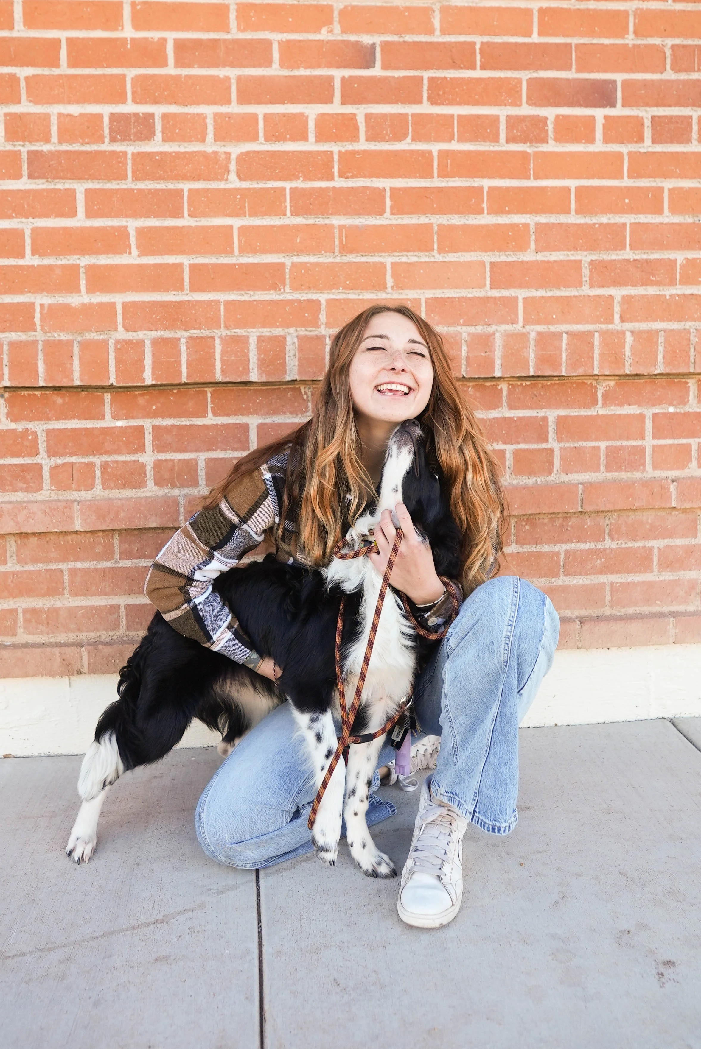 Young woman with long, wavy hair happily hugging a black and white dog against a brick wall.