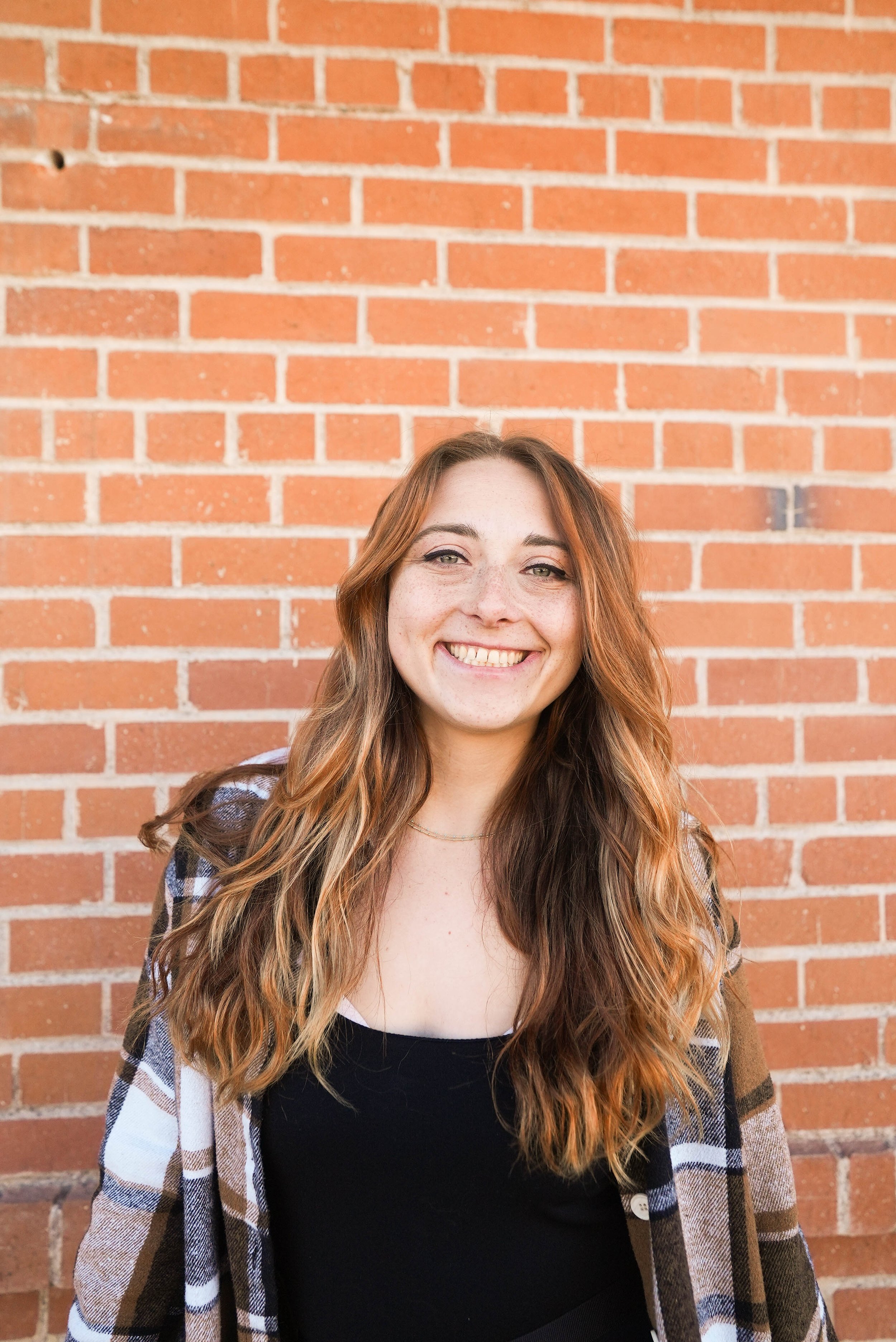 A young woman with long wavy red hair smiling in front of a brick wall.