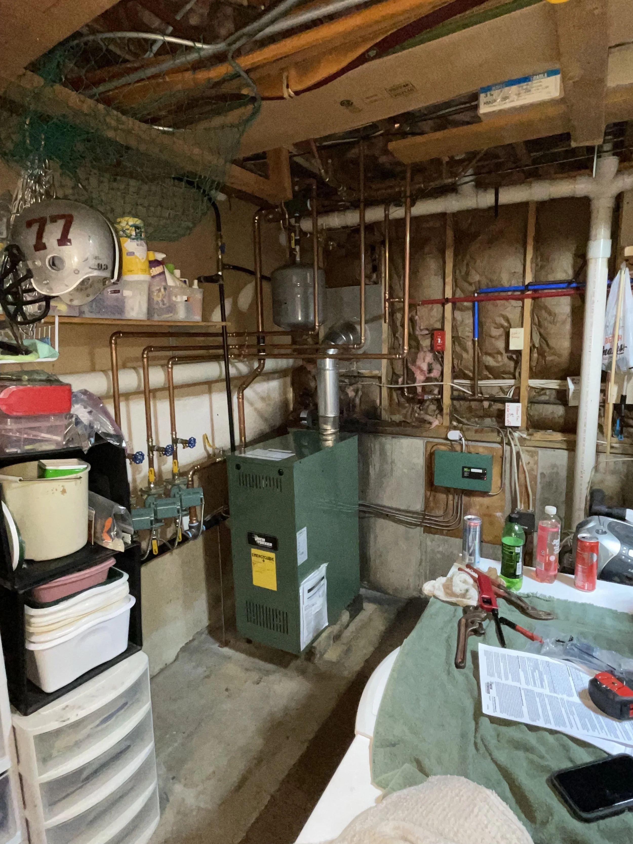 A cluttered basement with a green furnace, copper piping, shelves with storage bins, a table with drinks, and various tools and objects scattered around.