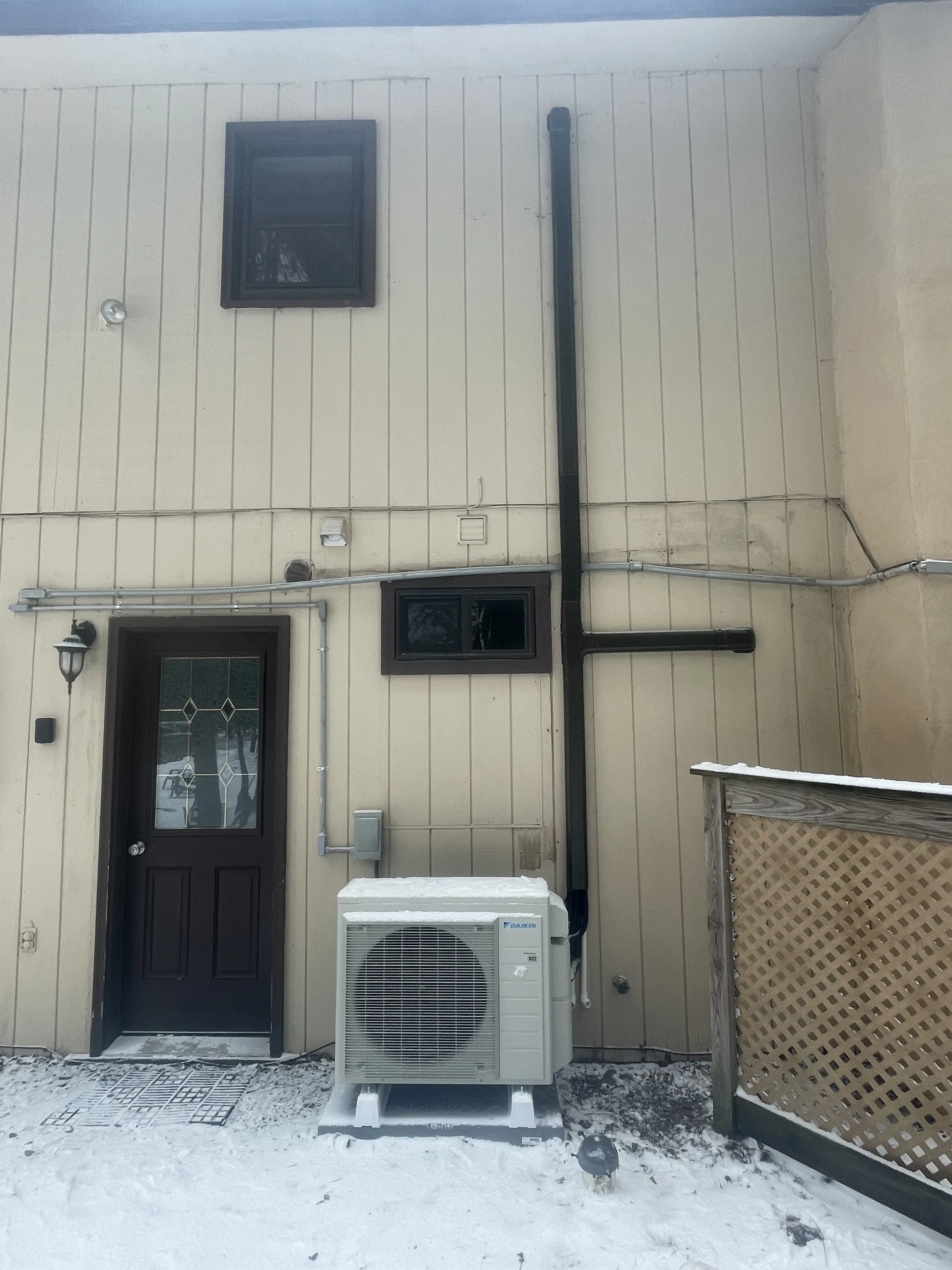 Back of house with beige siding, black door with decorative glass, small upper and lower windows, outdoor heating and cooling unit, and snow-covered ground.