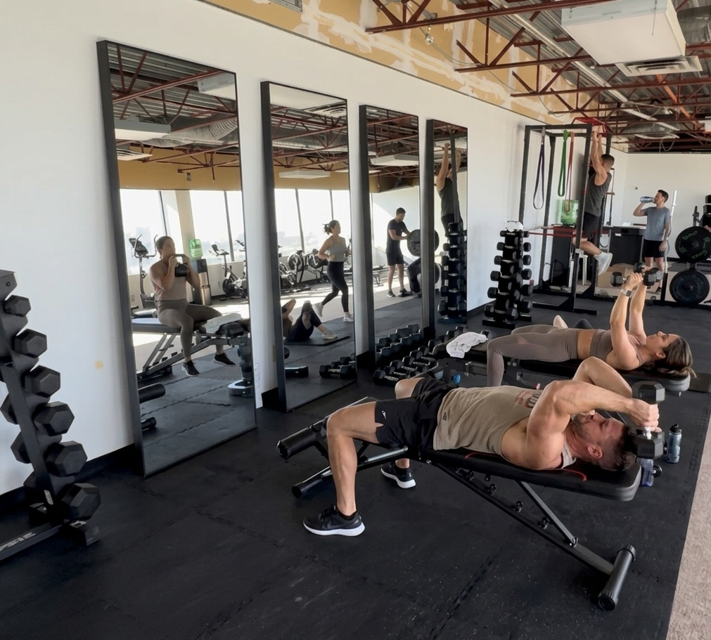 People working out in a gym, including a man lying on a bench doing a dumbbell exercise and a woman on a mat stretching or doing a workout. Mirrors reflect the gym equipment and other individuals exercising, with fitness bikes and weights visible.